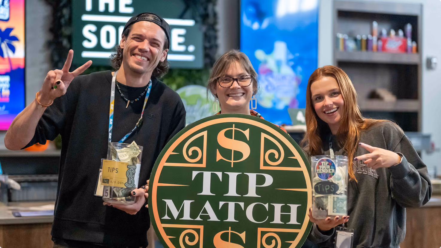 Three smiling employees at The Source Craft Cannabis Dispensary in Northwest Arkansas holding a green and gold, round ‘Tip Match' promotional sign and tip jars, showcasing community collaboration and positive workplace culture in a modern medical marijuana dispensary setting.