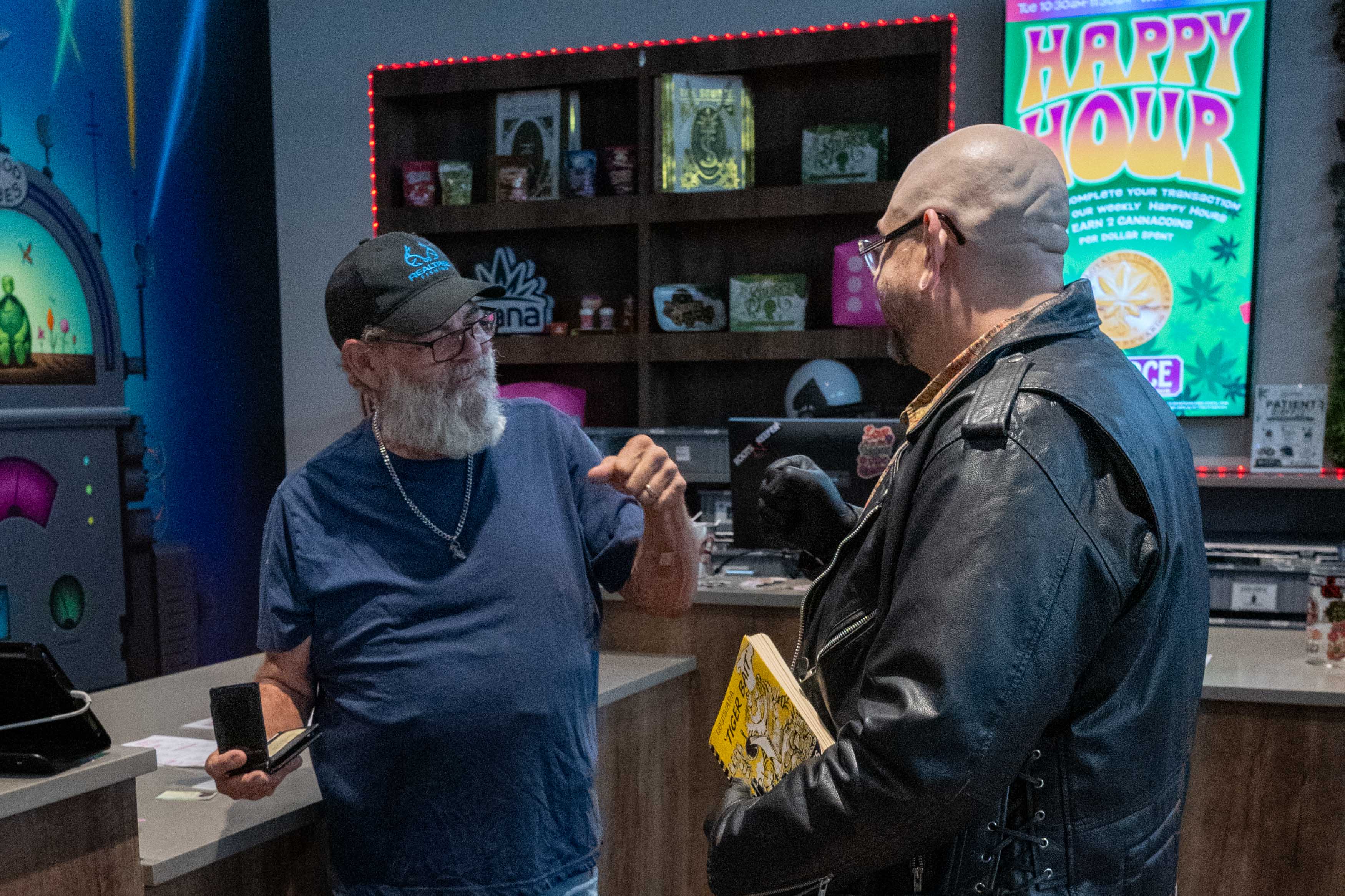Budtender Garrick in conversation with the same patient, who holds the 'Tiger Bait' cookbook gift, near the register on The Source's salesfloor, with a 'Happy Hour' promotional screen visible behind them."