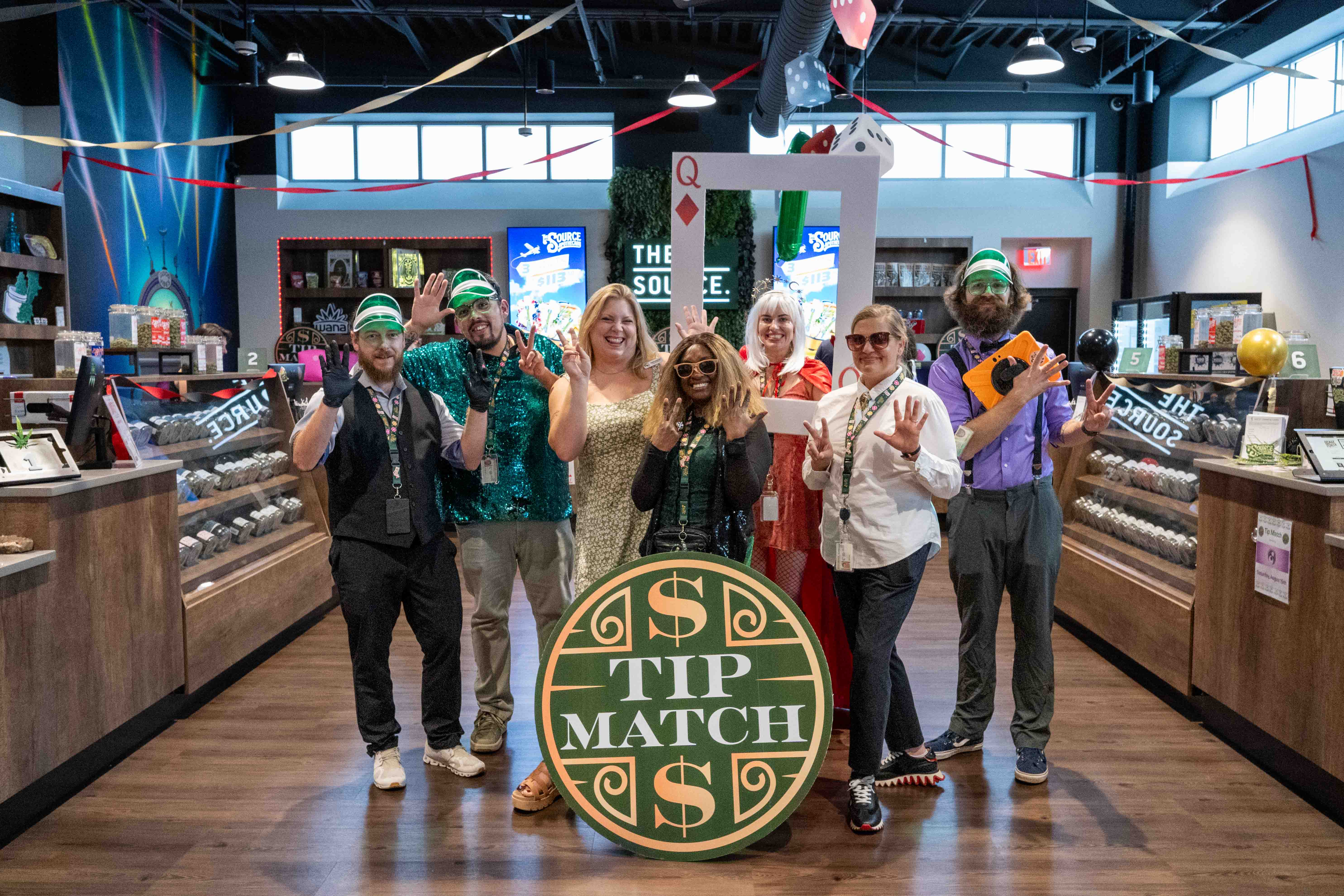 Tip Match with NWA Women's Shelter at The Source's Lucky 7th Anniversary event: seven team members in casino-themed costumes, including a dealer vest and a giant playing card photo frame, posing together in the retail aisle holding the round green and gold Tip Match sign, with dice-shaped decorations hanging overhead.
