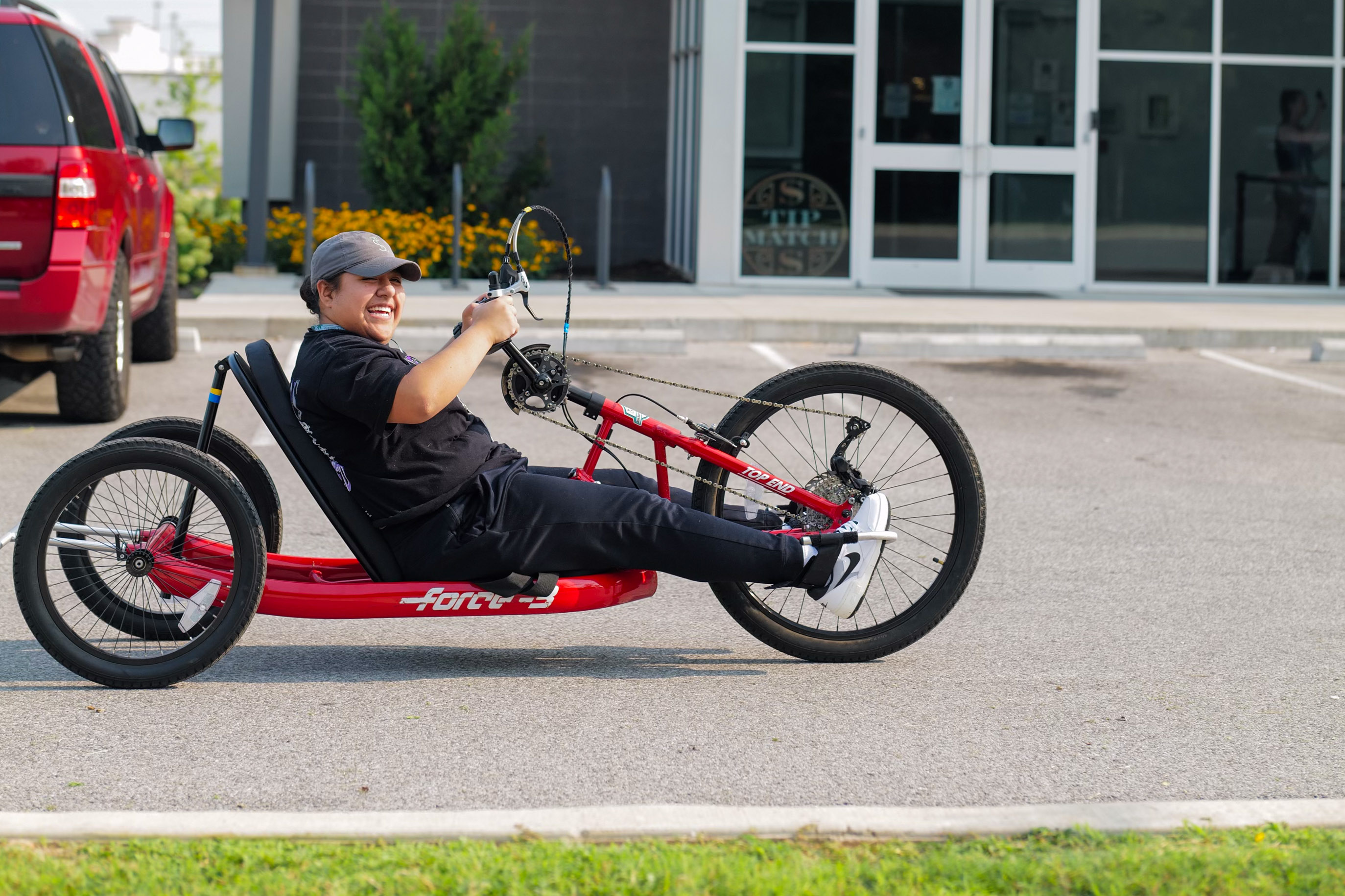 A member of The Source team riding a red Force-3 recumbent handcycle in The Source’s parking lot, smiling while wearing black clothing and gray cap, adaptive sports equipment for parasports and inclusive cycling.