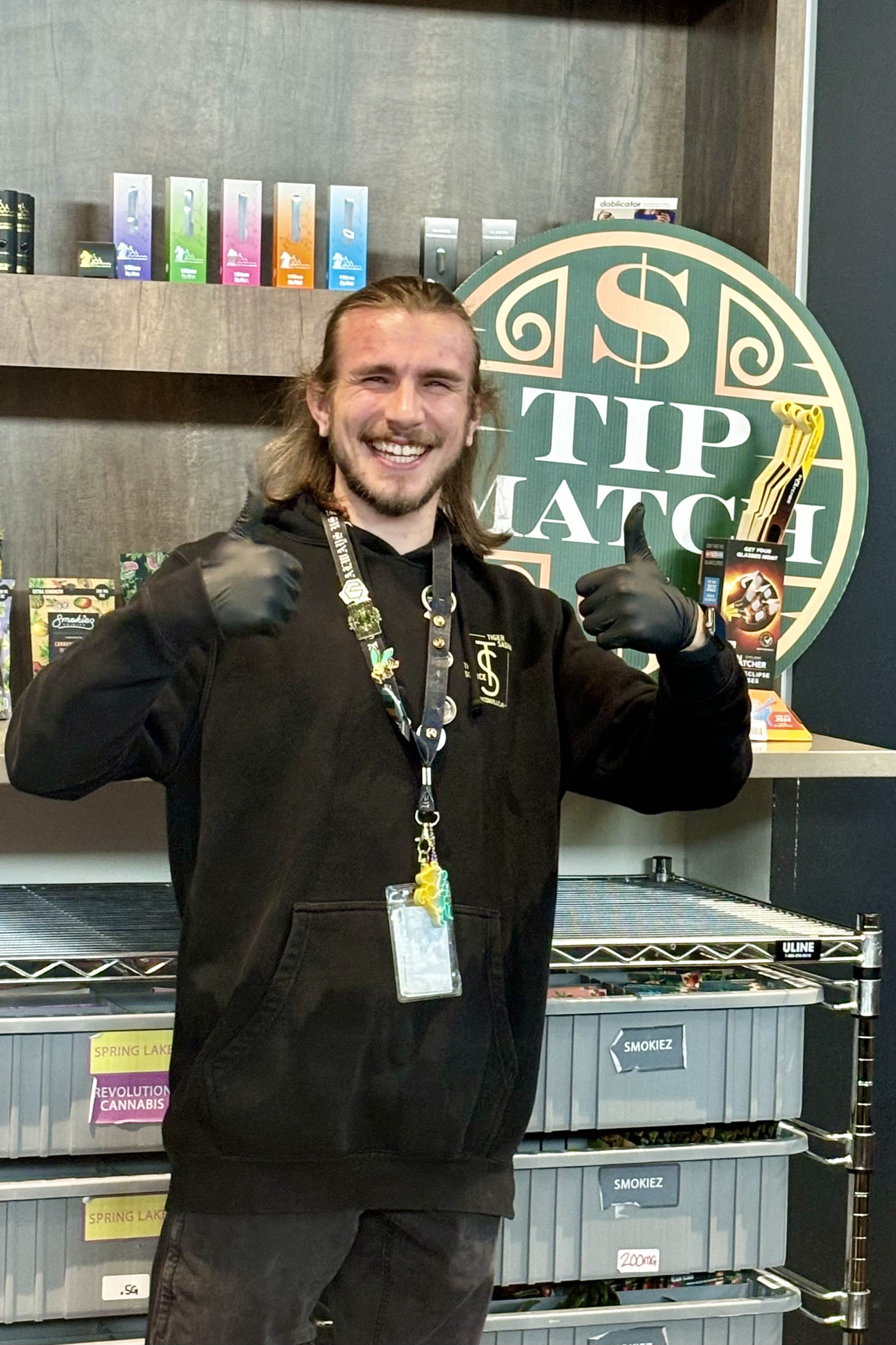 A smiling budtender in a black hoodie giving two thumbs up at The Source Dispensary. He is standing in front of retail shelving with colorful products and a round “Tip Match” sign.
