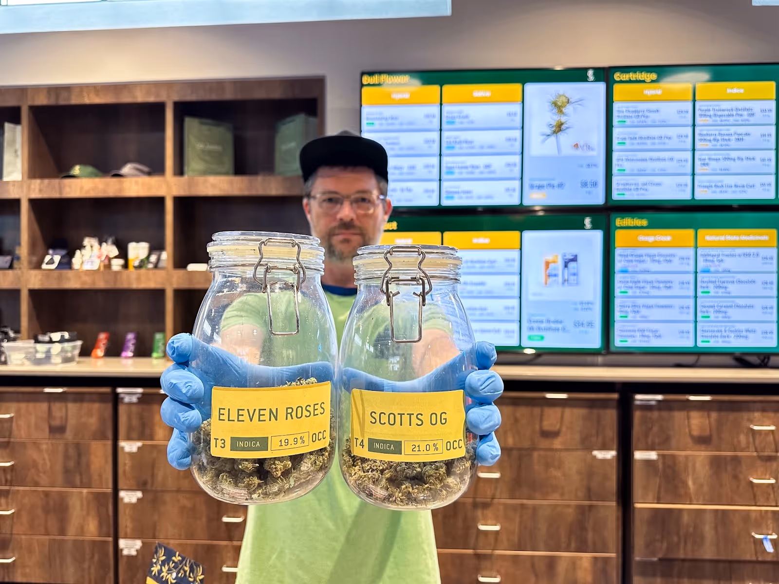 Budtender at The Source cannabis dispensary located in Rogers, Arkansas, holds up jars of popular cannabis indica flower strains, Scott’s OG and Eleven Roses by Osage Creek Cultivation.