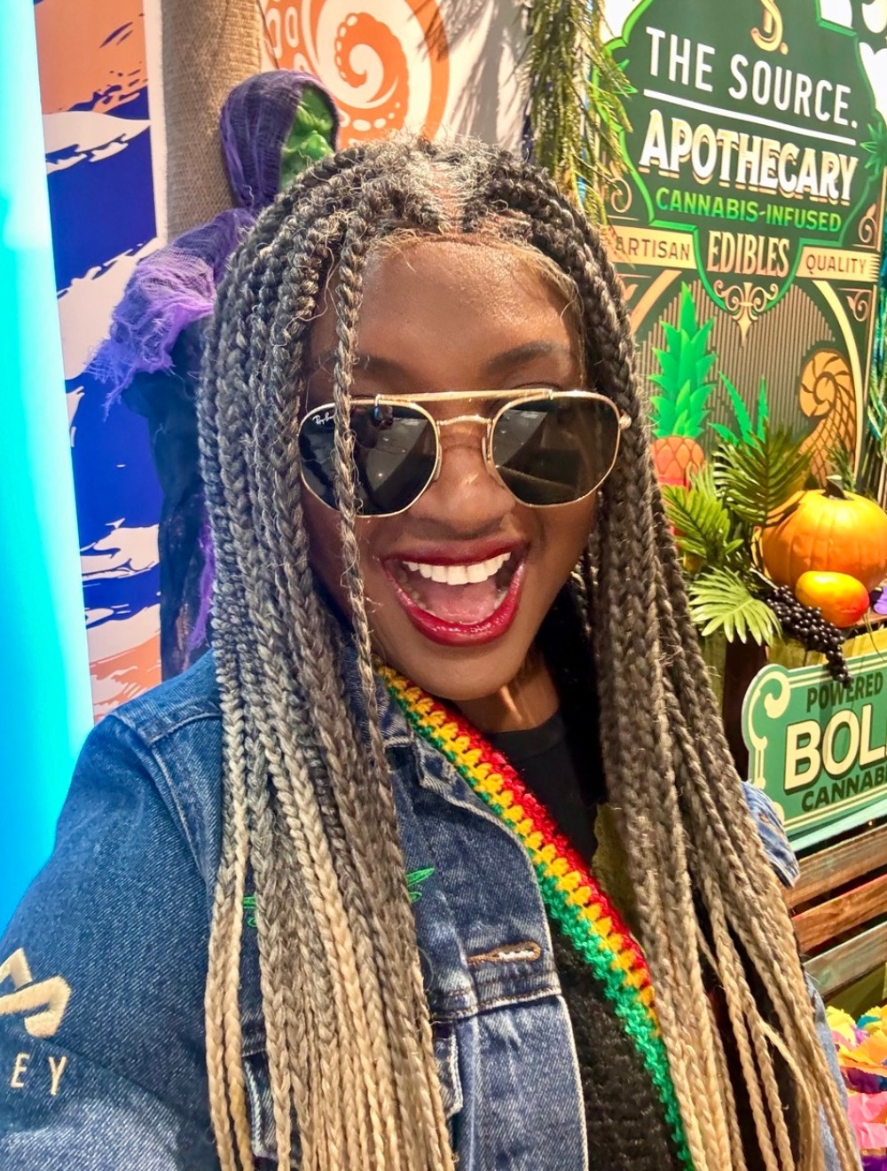 Rochelle Bradshaw, cannabis brand ambassador for The Source dispensary in Rogers, Arkansas smiling with The Source Apothecary signage and tropical decor.
