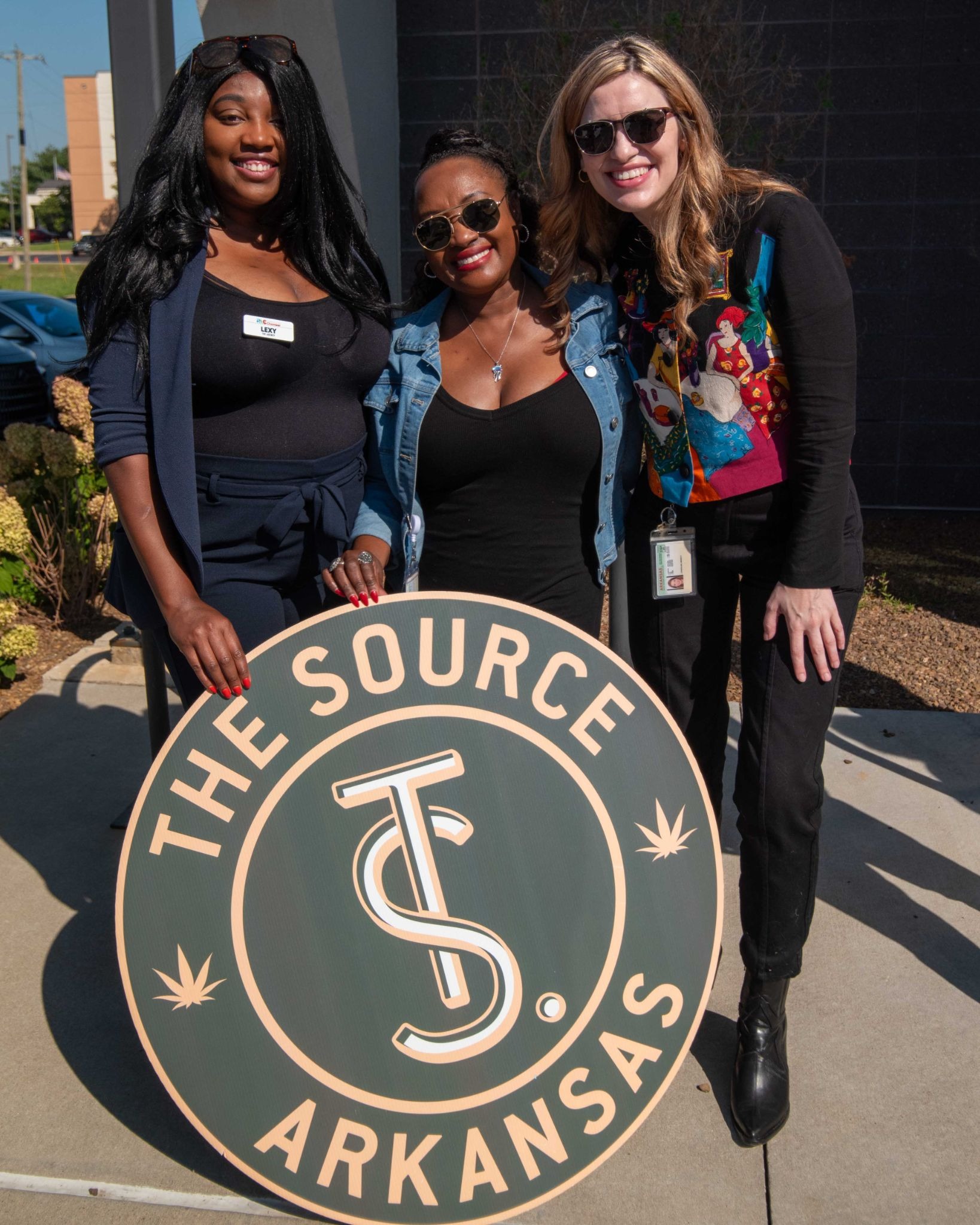 The Source logo featuring stylized 'TS' design on branded sign being held by Rogers Lowell Chamber of Commerce members at The Source in NWA.