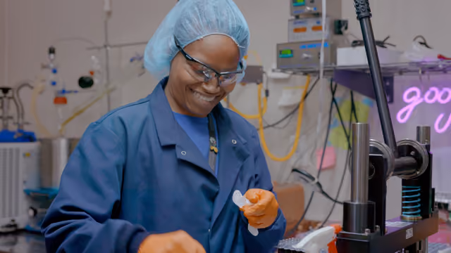Cannabis lab technician in blue scrubs and hairnet examining product sample at Arkansas medical marijuana processing facility.