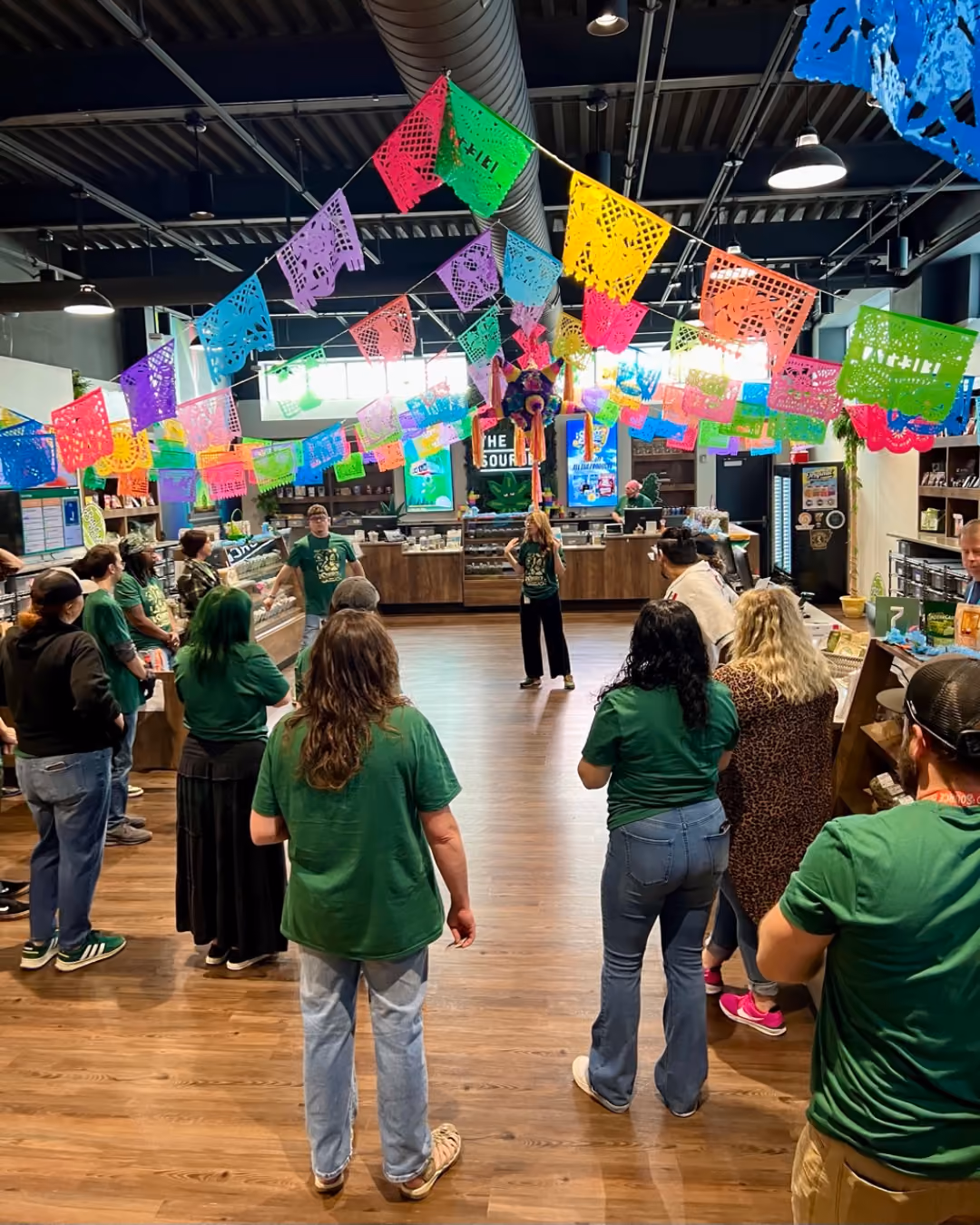 The Source staff in green shirts gathered inside dispensary decorated with colorful papel picado banners for 4/20 Fiesta celebration.
