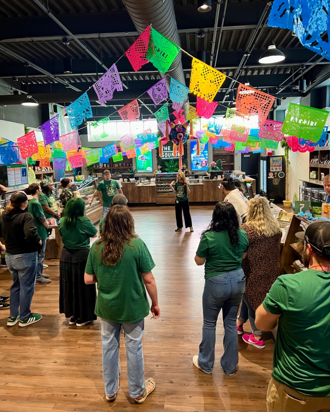 The Source staff in green shirts gathered inside dispensary decorated with colorful papel picado banners for 4/20 Fiesta celebration.
