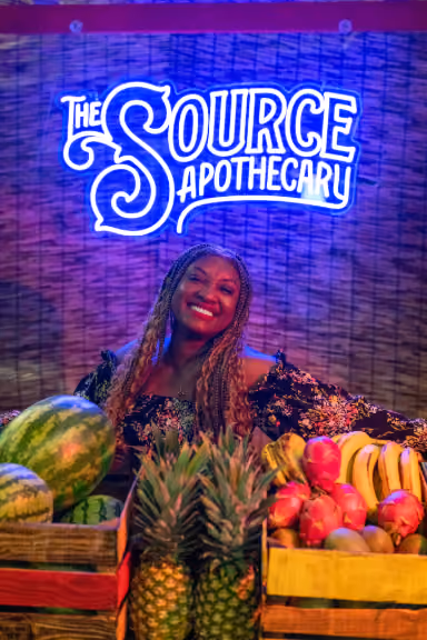 Rochelle Bradshaw smiling beneath The Source Apothecary neon sign surrounded by tropical fruit, celebrating the Island Time product line at The Source Dispensary in Rogers, Arkansas.