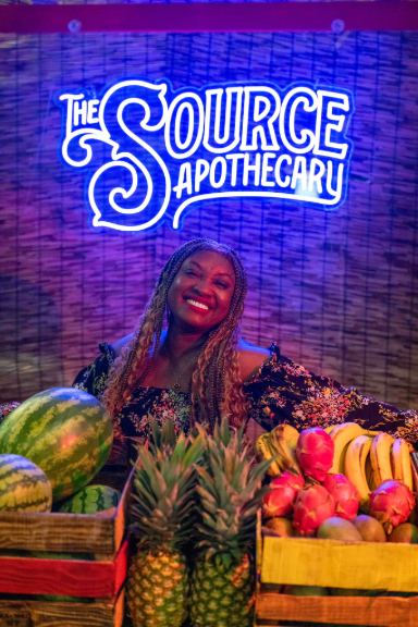 Rochelle Bradshaw smiling beneath The Source Apothecary neon sign surrounded by tropical fruit, celebrating the Island Time product line at The Source Dispensary in Rogers, Arkansas.