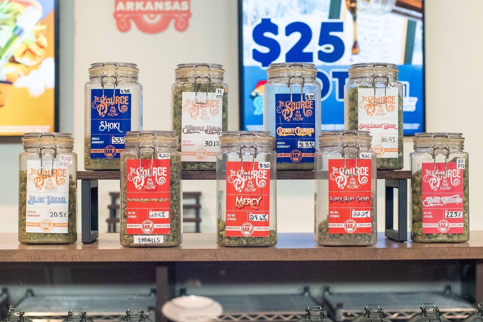 In The Flow deli-style cannabis flower jars on display at The Source Dispensary in Rogers, Arkansas, showing strains including Shoki, Truffle Hound, Donny Burger, Strawberry Mentz, Lilac Diesel, Platinum Lemon Cherry Gelato, Mercy, Super Buff Cherry, and White Wedding, with handwritten price tags ranging from $20 to $35.