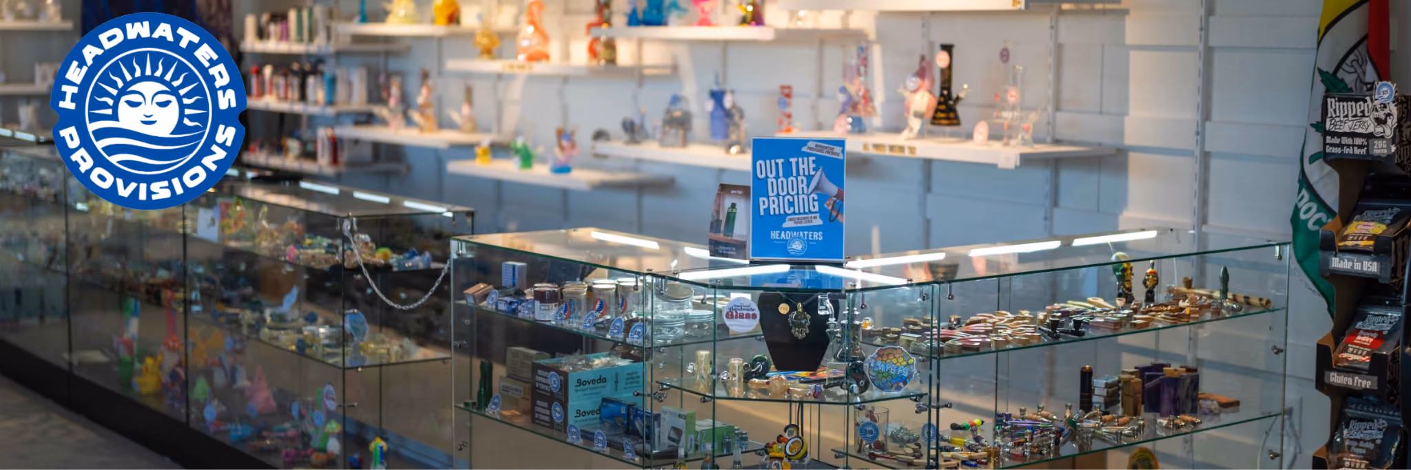 Headwaters Provisions logo over glass display cases of glass pipes, bongs, and smoking accessories, with an 'Out the Door Pricing' sign on the counter.