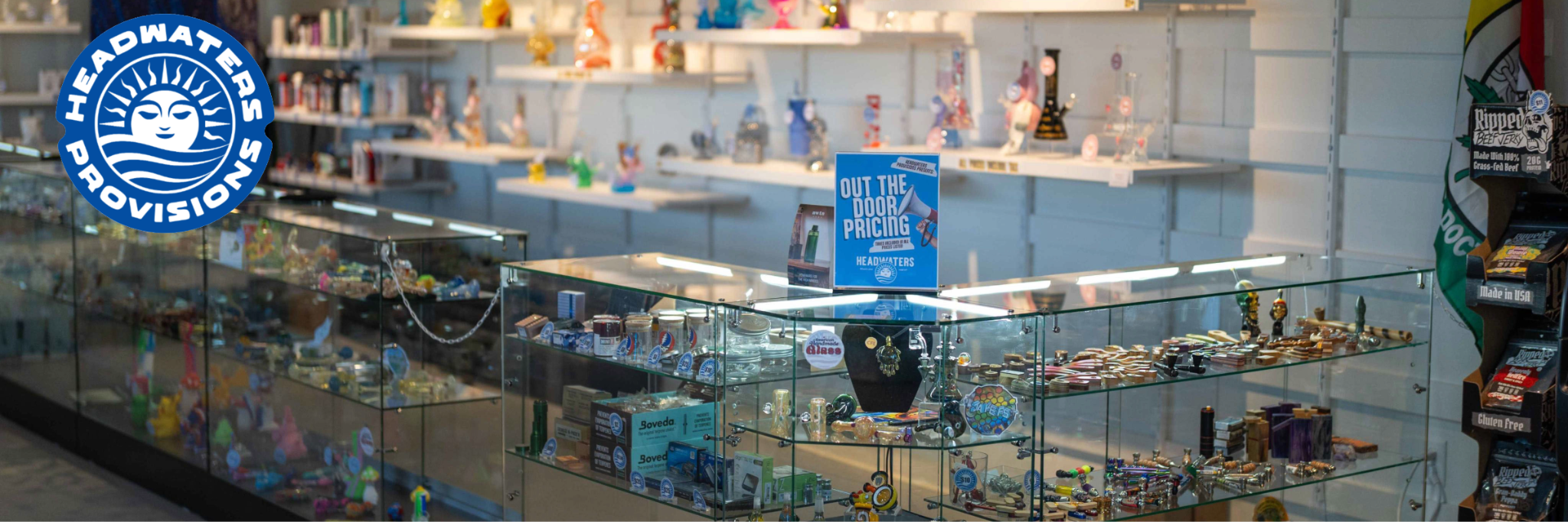 Headwaters Provisions logo over glass display cases of glass pipes, bongs, and smoking accessories, with an 'Out the Door Pricing' sign on the counter.