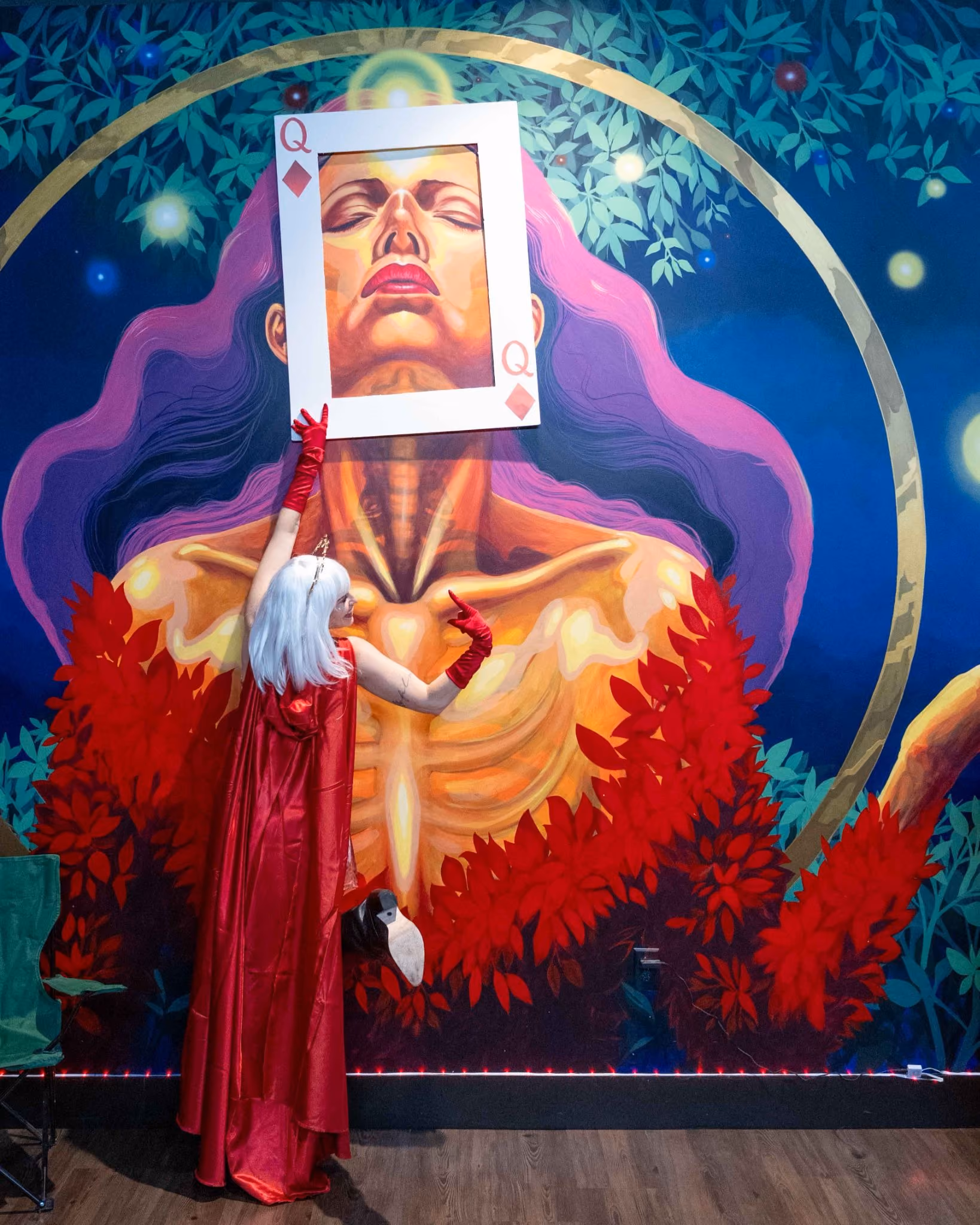 The Source's CEO, Amanda Strickland, in a red cape and crown poses as the Queen of Diamonds against a vivid mural, celebrating the female-led spirit of the Lucky 7 Anniversary.