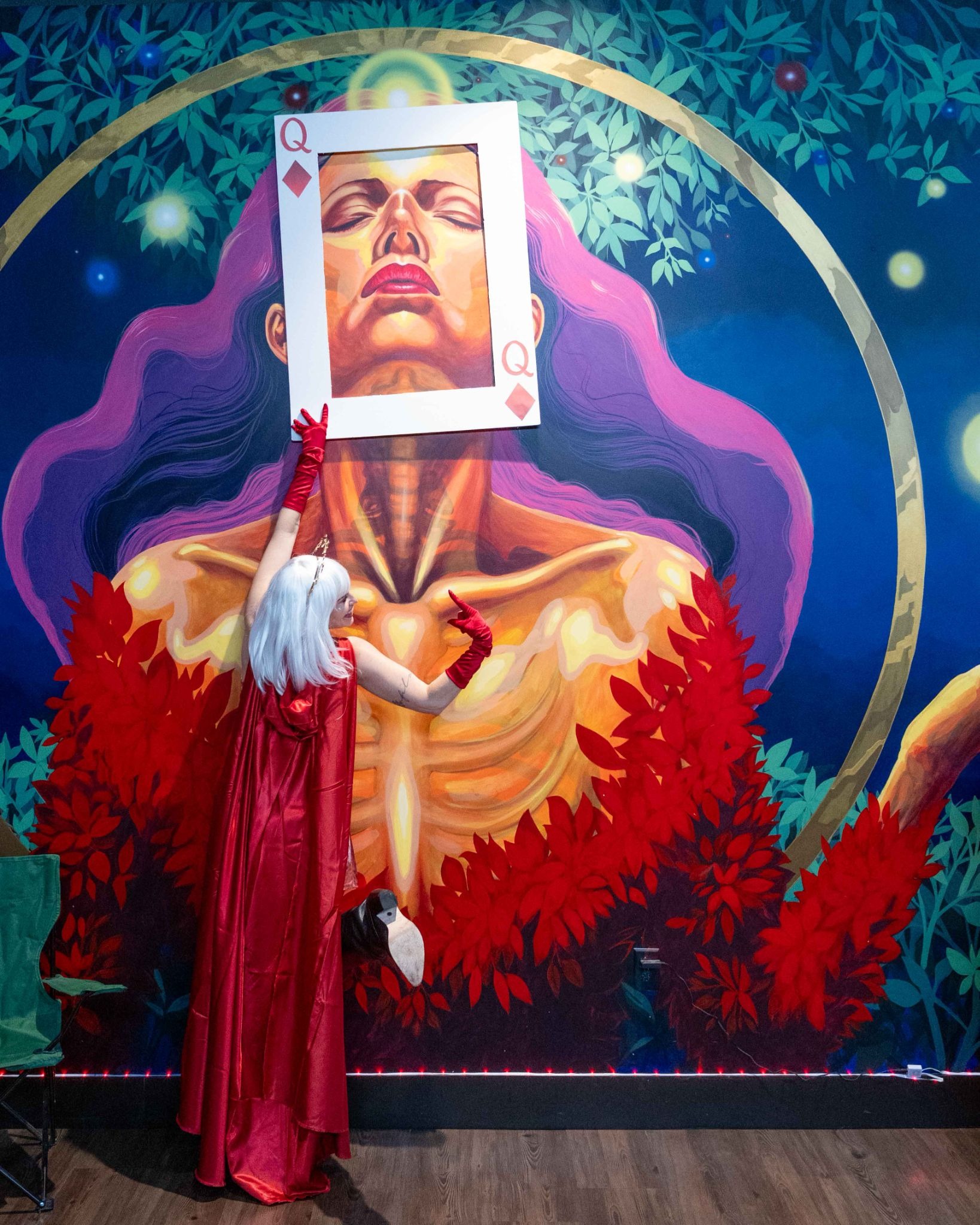 The Source's CEO, Amanda Strickland, in a red cape and crown poses as the Queen of Diamonds against a vivid mural, celebrating the female-led spirit of the Lucky 7 Anniversary.