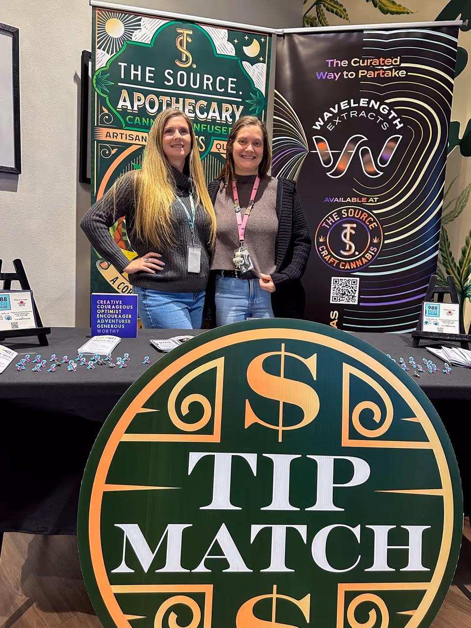 Two women wearing lanyards standing in front of cannabis retail displays at The Source dispensary. Behind them are decorative signs showing 'Cannabis-Infused' products and the company's logo with rainbow holographic effects.