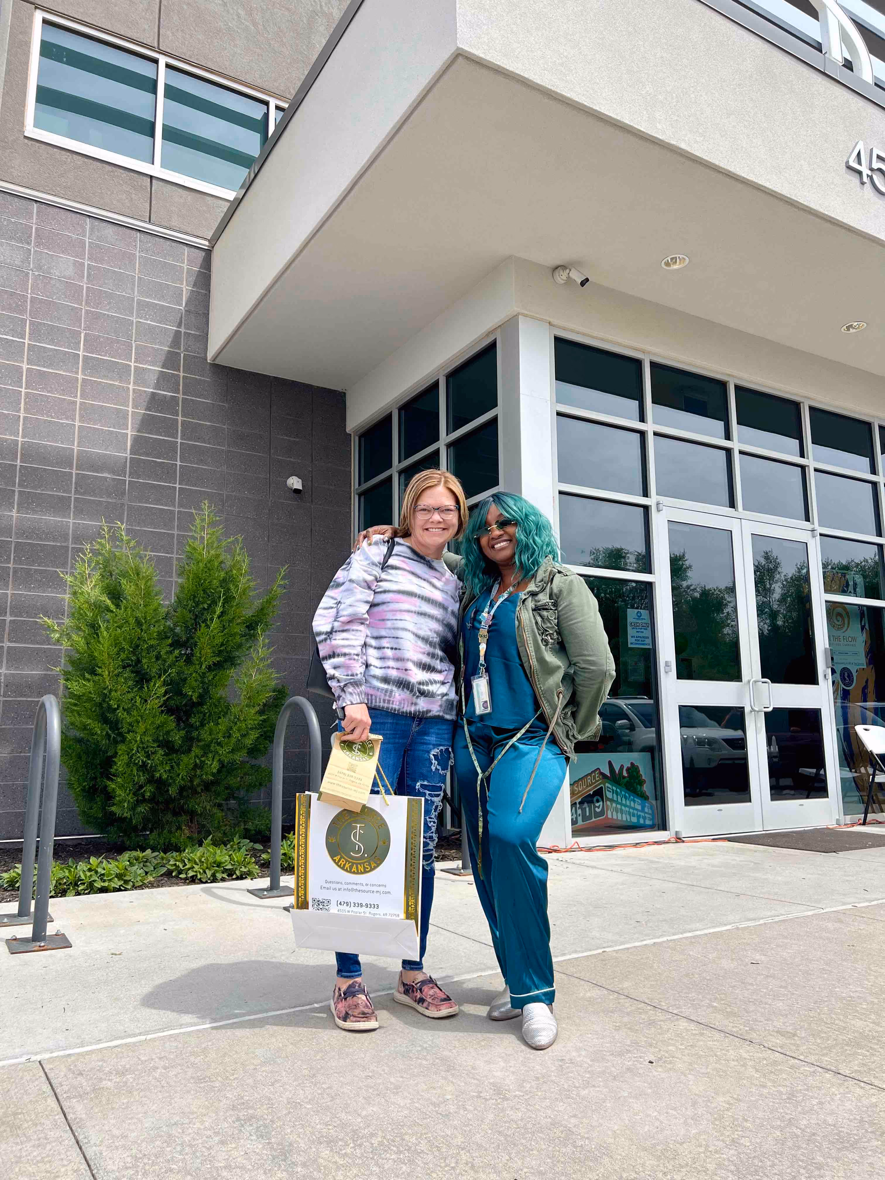 Two people standing outside a modern commercial building entrance, one wearing a tie-dye sweatshirt and holding a shopping bag, the other wearing a teal outfit with green hair and a military-style jacket. Evergreen shrubs near the entrance of The Source.