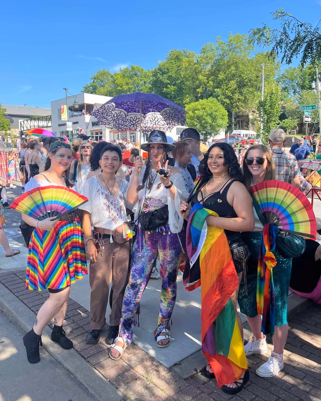 Tip Match with NWA Equality Inc. at the NWA Pride parade: five Source team members posing together on Dickson Street holding rainbow fans and a pride flag, with a decorative purple lace parasol and parade crowd visible behind them.