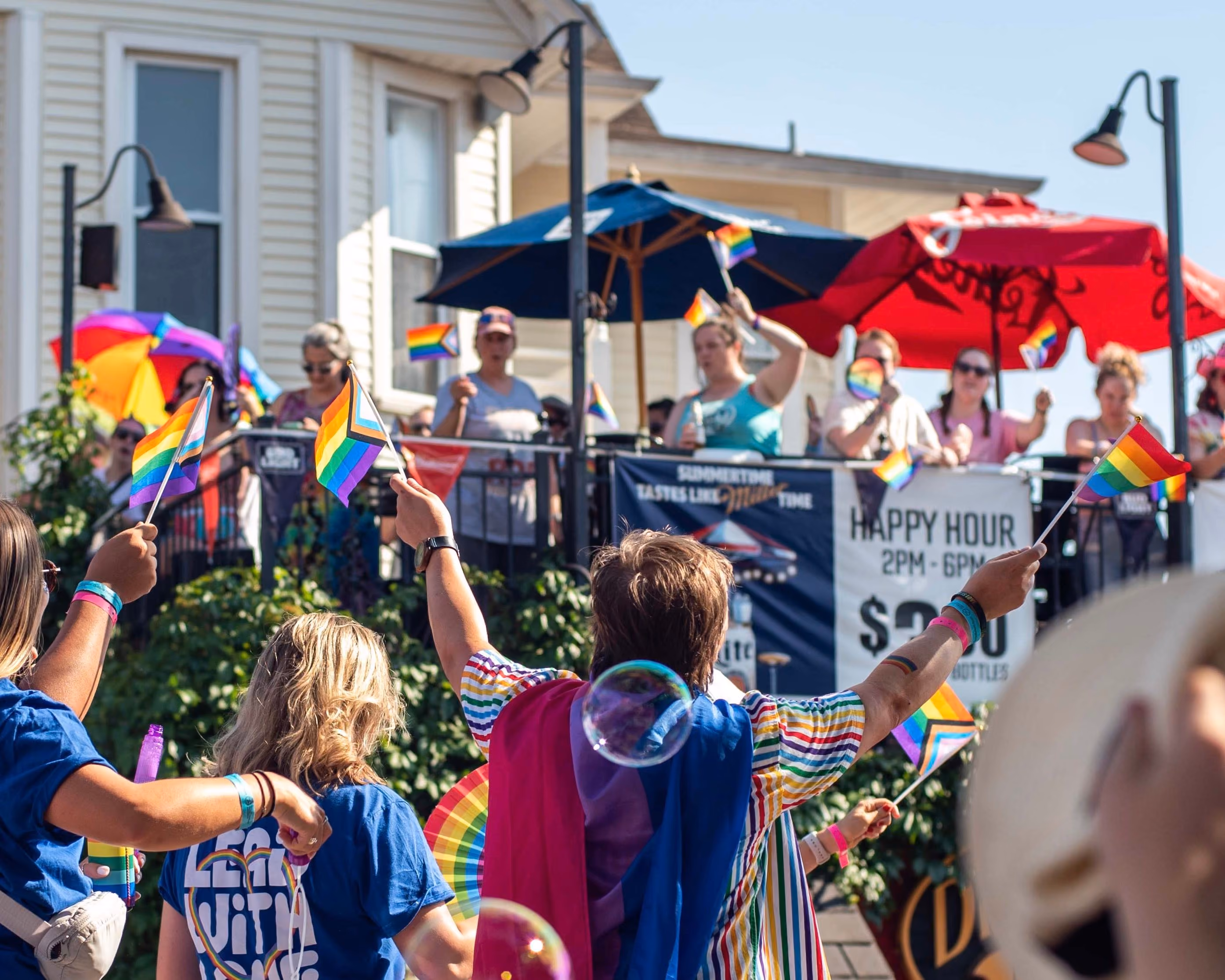 Pride celebration outside restaurant patio showing diverse crowd waving rainbow flags during happy hour, with participants wearing colorful pride-themed clothing and accessories. Outdoor venue features umbrellas and happy hour signage.