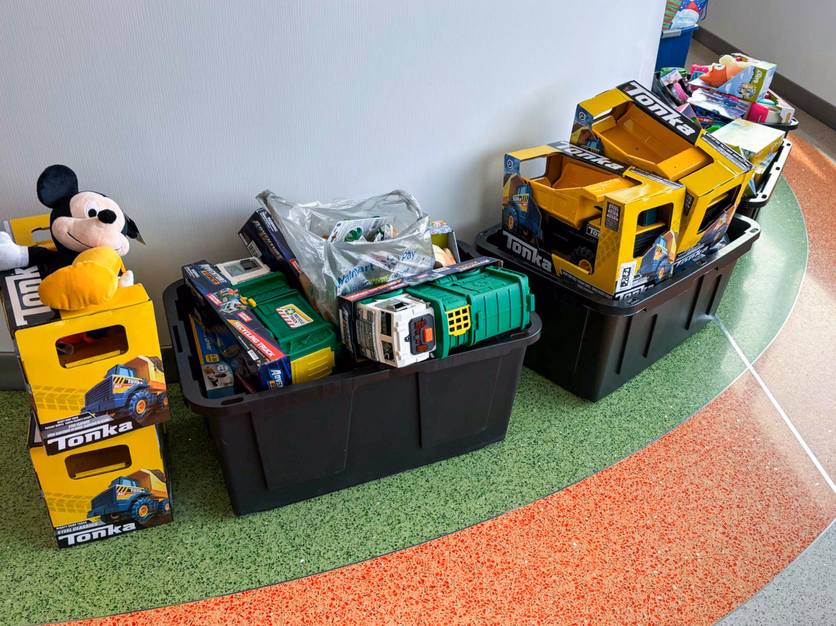 Toy donations collected for The Source's Holiday Toy Drive benefiting Arkansas Children's Hospital, with Tonka dump trucks, a Mickey Mouse plush, and other toys overflowing from storage totes.