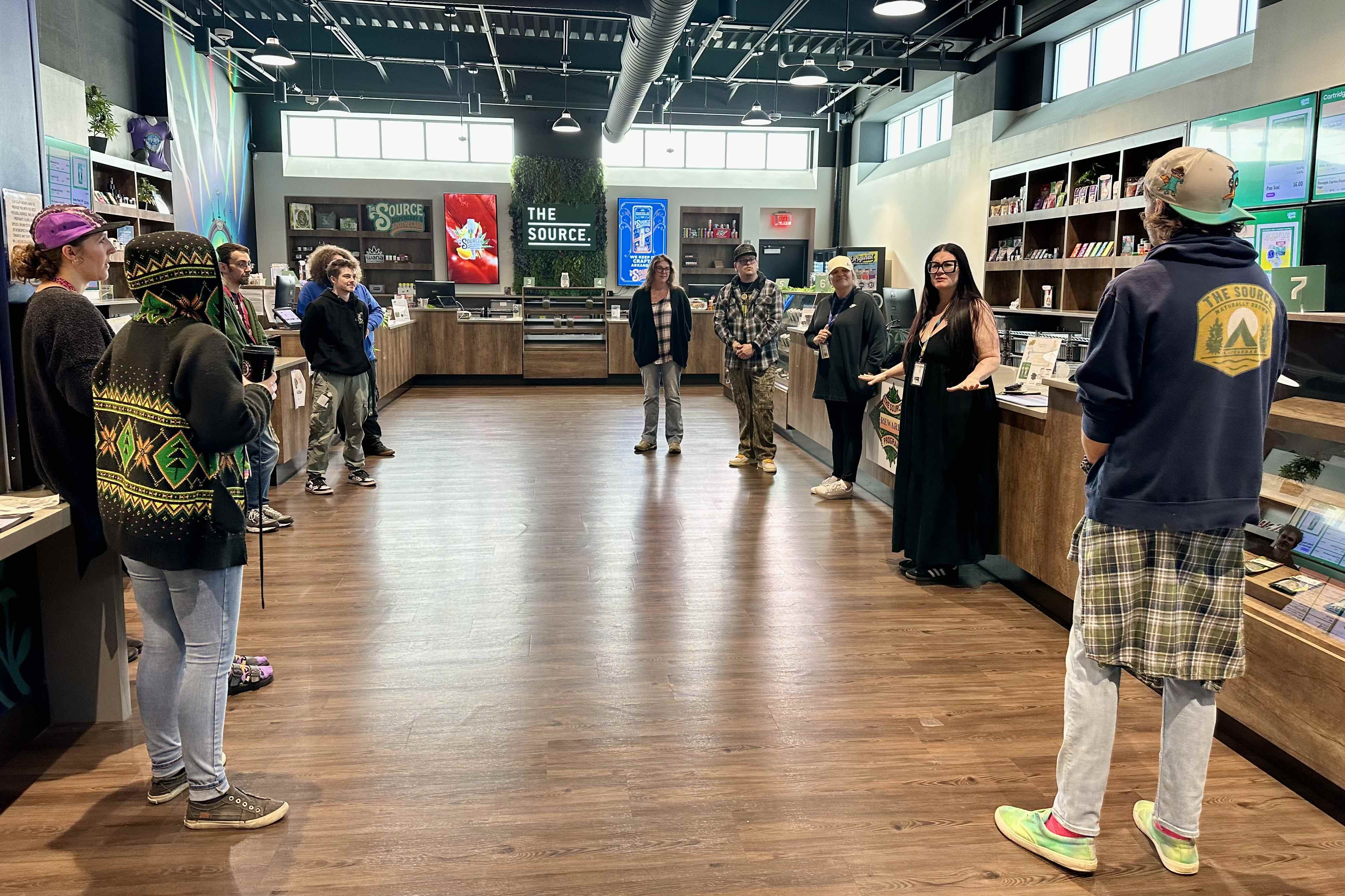 Interior of The Source cannabis dispensary showing staff members having a meeting in a modern retail space with wood floors, branded signage, display cases, and industrial ceiling. The well-lit store features clean design elements with wooden counters.