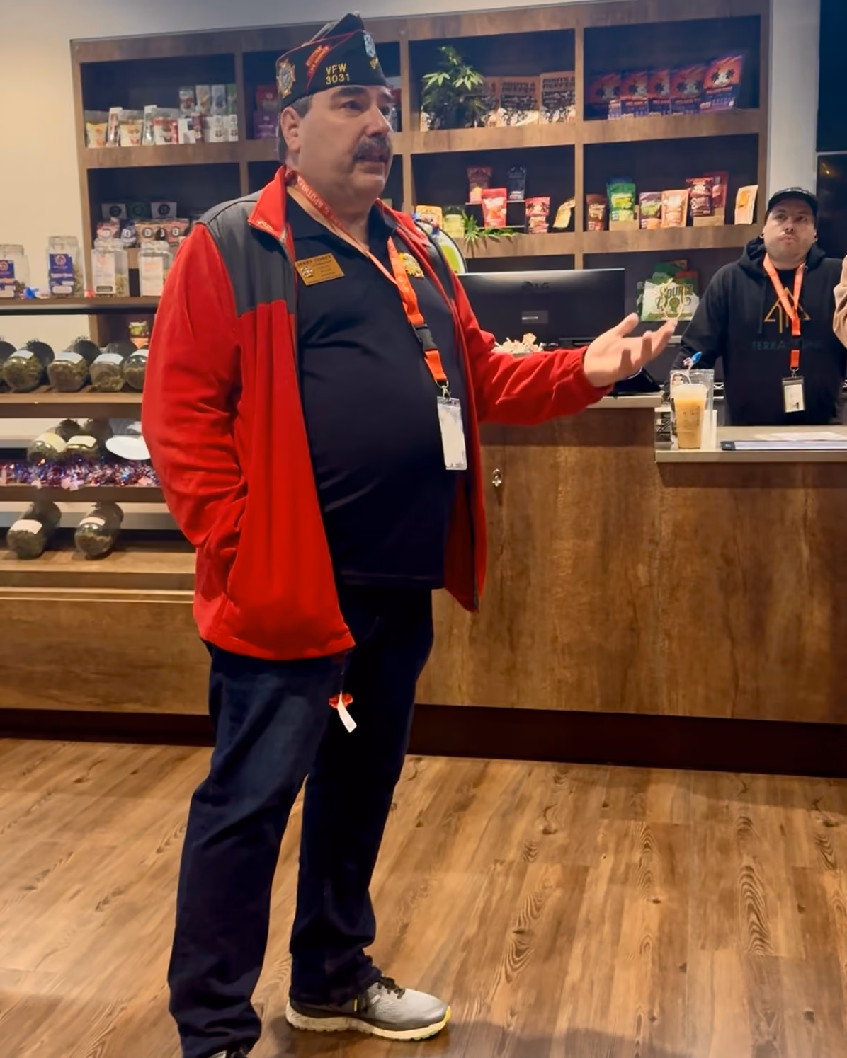 Tip Match with VFW Post 3031 at The Source Dispensary & Apothecary: a veteran wearing a red jacket and VFW garrison cap reading 'VFW 3031,' gesturing while speaking in front of the retail display shelves, with a staff member visible behind the counter.