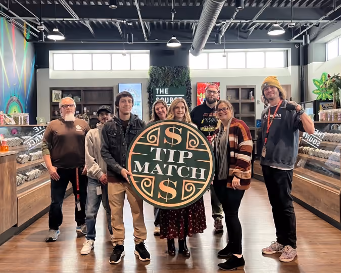 Tip Match with NWA Crisis Intervention Center at The Source Dispensary & Apothecary: seven smiling team members standing together in the retail showroom holding the round green and gold Tip Match sign, with product display cases and 'THE SOURCE' signage