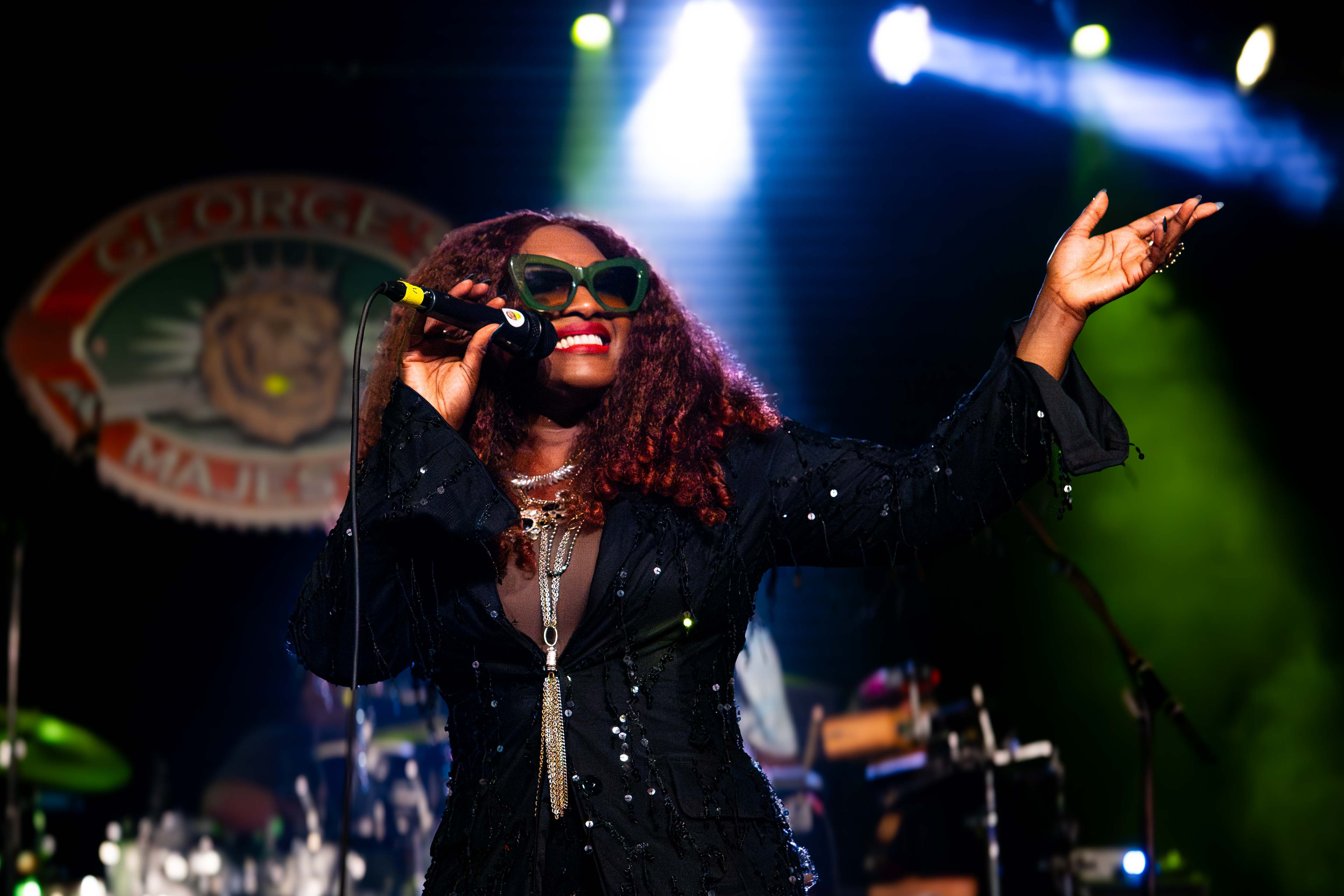 Rochelle Bradshaw performing on stage in green cat-eye sunglasses and a sequined black outfit, arm raised mid-performance, with George's Majestic Lounge's lion logo glowing behind her under blue and green stage lighting.