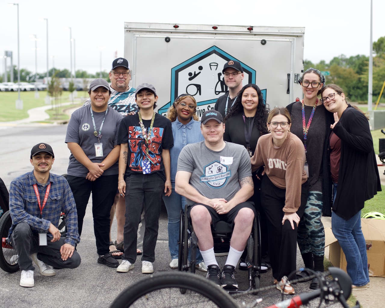 Tip Match with Ozark Adaptive Sports Association at The Source: a group of eleven staff members and OZASA representatives posing outdoors in front of the organization's equipment trailer, with an adaptive cycling wheelchair visible in the foreground.
