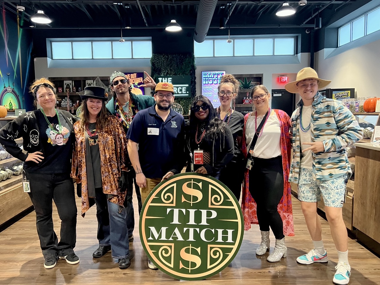 Tip Match with For The Love NWA at The Source Dispensary & Apothecary: eight team members in Halloween costumes posing together in the retail showroom holding the round green and gold Tip Match sign, with 'THE SOURCE' neon signage and pumpkin decor visible