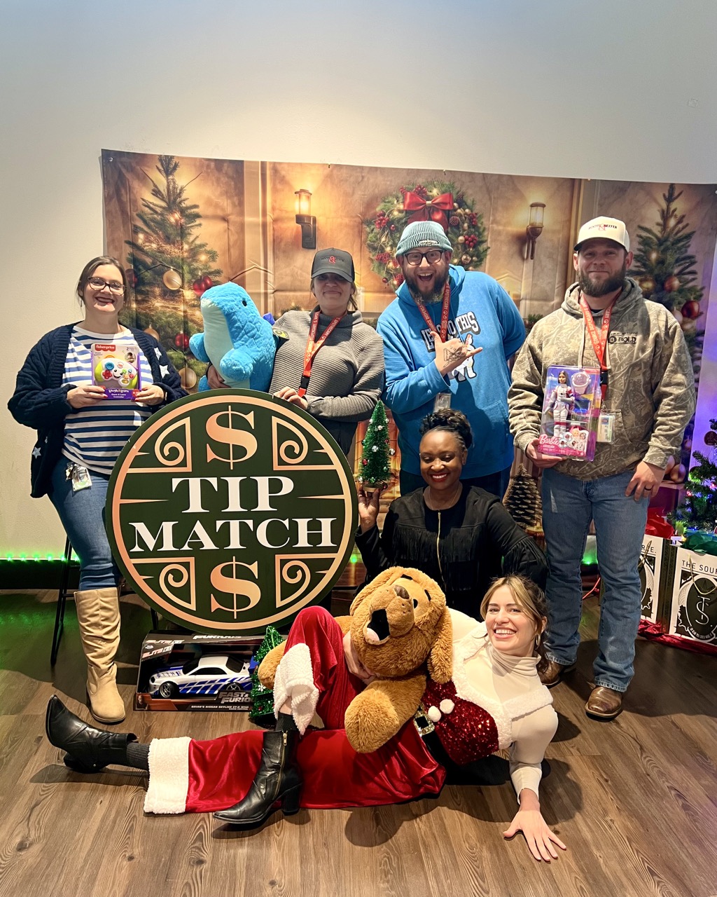 Tip Match for the Holiday Toy Drive at The Source Dispensary & Apothecary: six team members in festive attire, including a Santa costume, posing together holding donated toys and a large plush dog beside the round green and gold Tip Match sign.