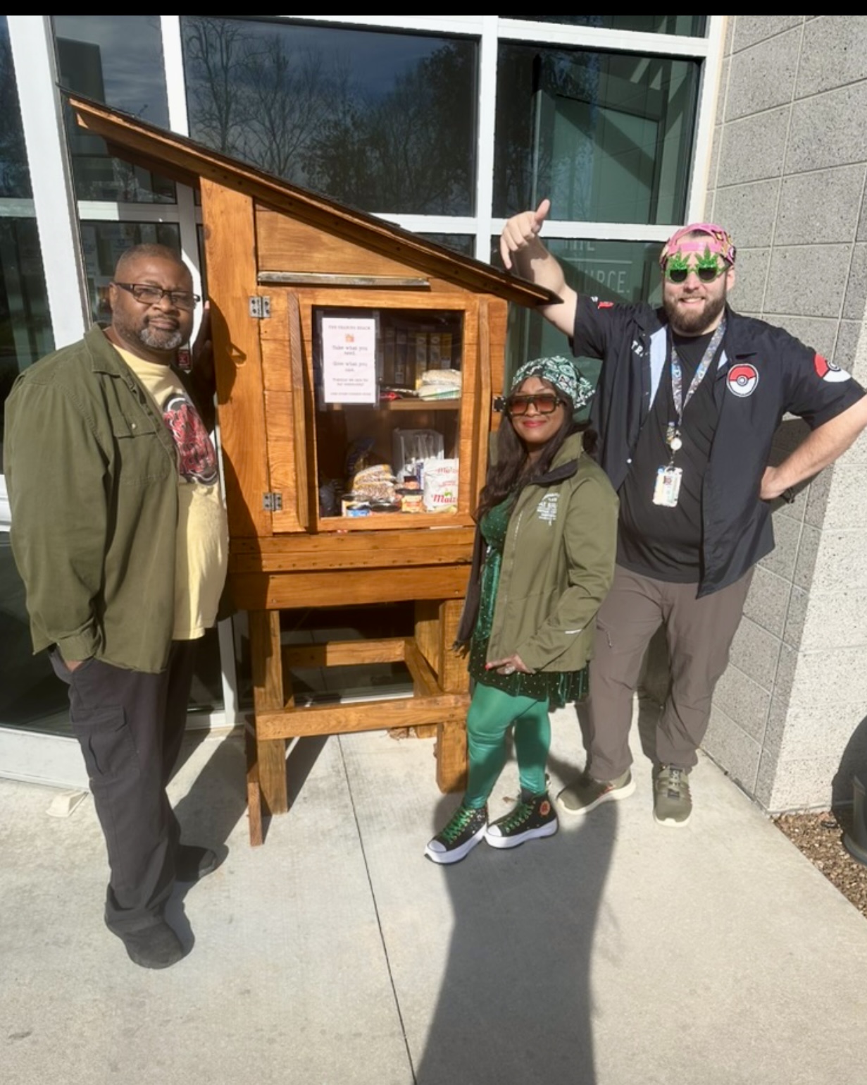 Nate Walls and two Source team members standing beside the Sharing Shack, stocked with canned goods and snacks outside The Source, with a 'take what you need, give what you can' sign posted on the door.