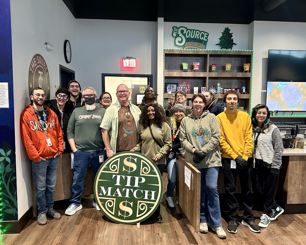 Tip Match with NWA Crisis Intervention Center at The Source Dispensary & Apothecary: fourteen team members in holiday sweaters and costumes, including one dressed as Scooby-Doo, posing together in the retail showroom holding the round green Tip match logo