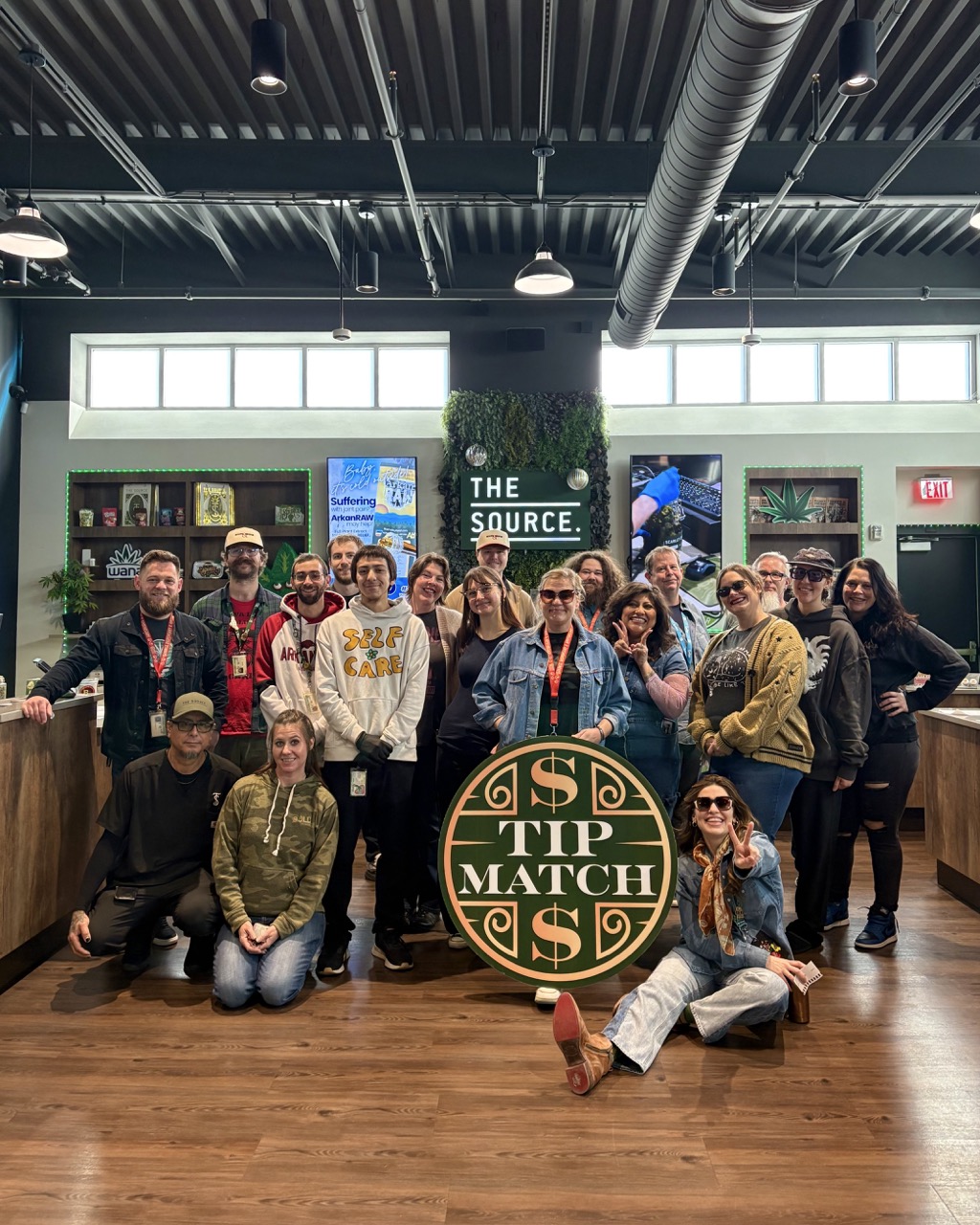 Tip Match with Bob & Rita Marley Foundation at The Source Dispensary & Apothecary: a large group of team members posing together in the retail showroom holding the round green and gold Tip Match sign, with 'THE SOURCE' signage and a living plant wall.
