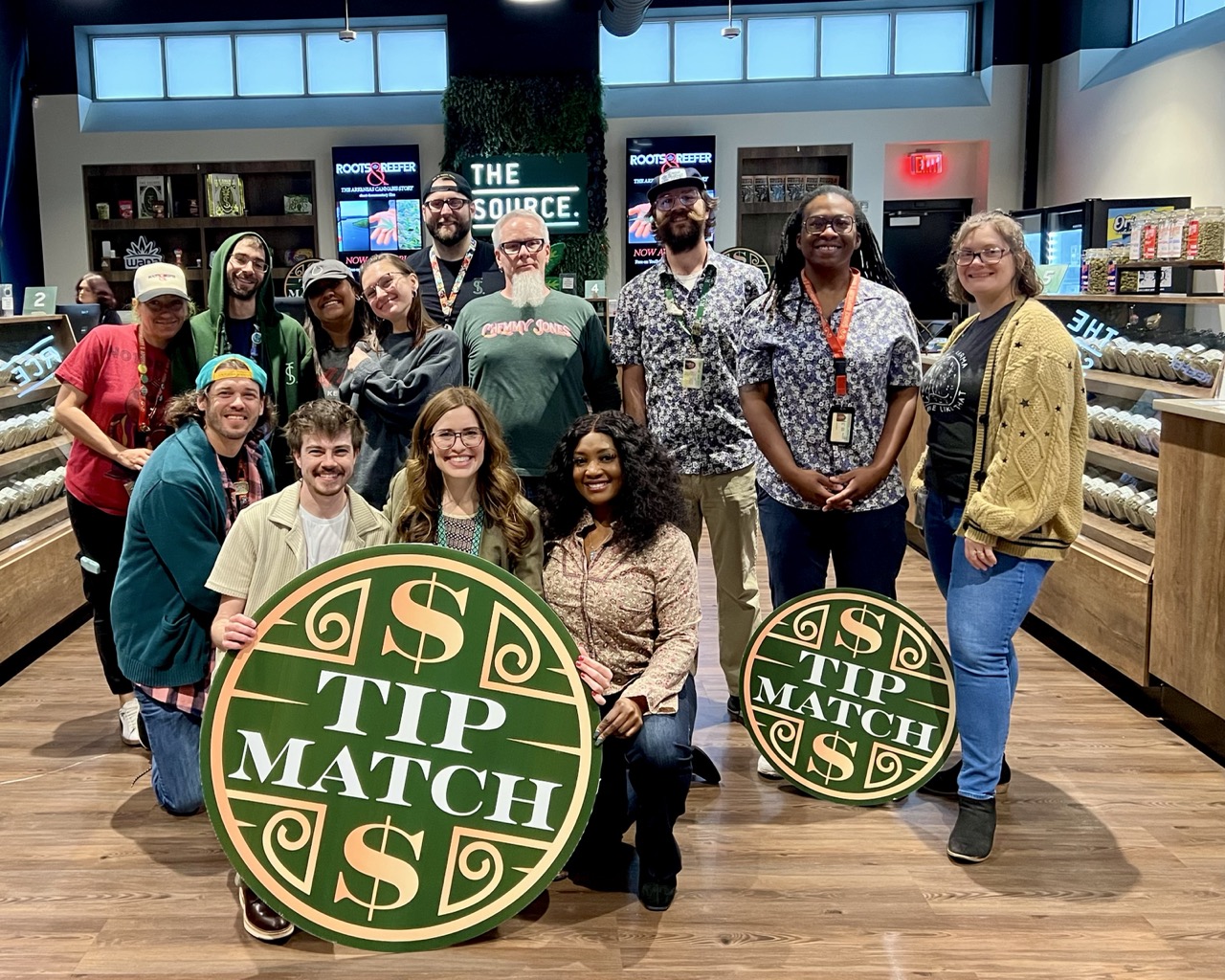 Tip Match with Women's Foundation of Arkansas at The Source Dispensary & Apothecary: eleven team members posing together in the retail showroom holding two round green and gold Tip Match signs, with 'THE SOURCE' signage and product display cases visible