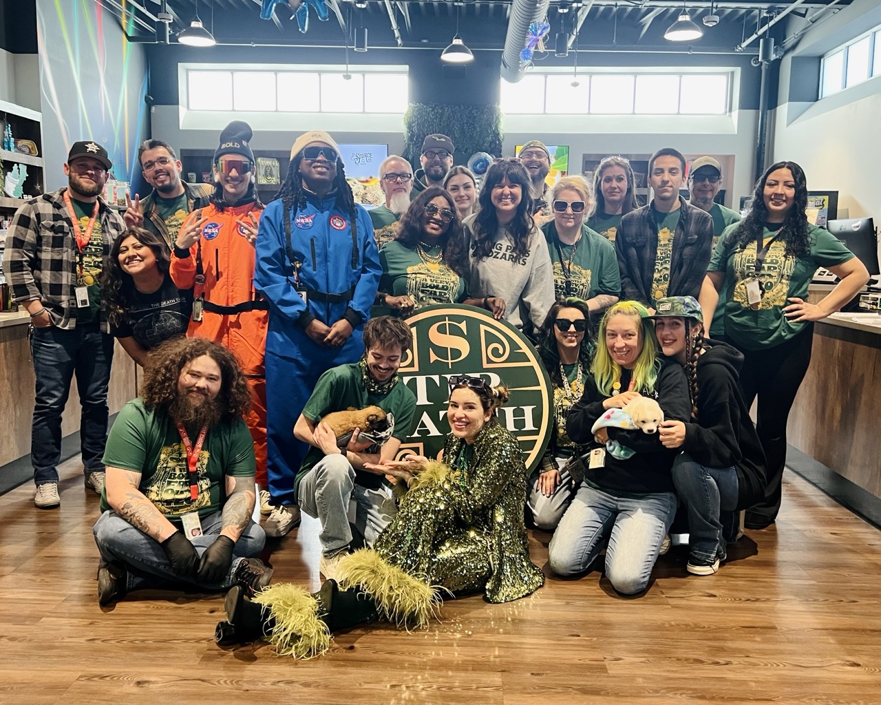  Large group photo for Tip Match with Big Paws of the Ozarks: team members in matching green shirts and costumes, including two dressed as astronauts, posing together holding puppies beside the round green and gold Tip Match sign.