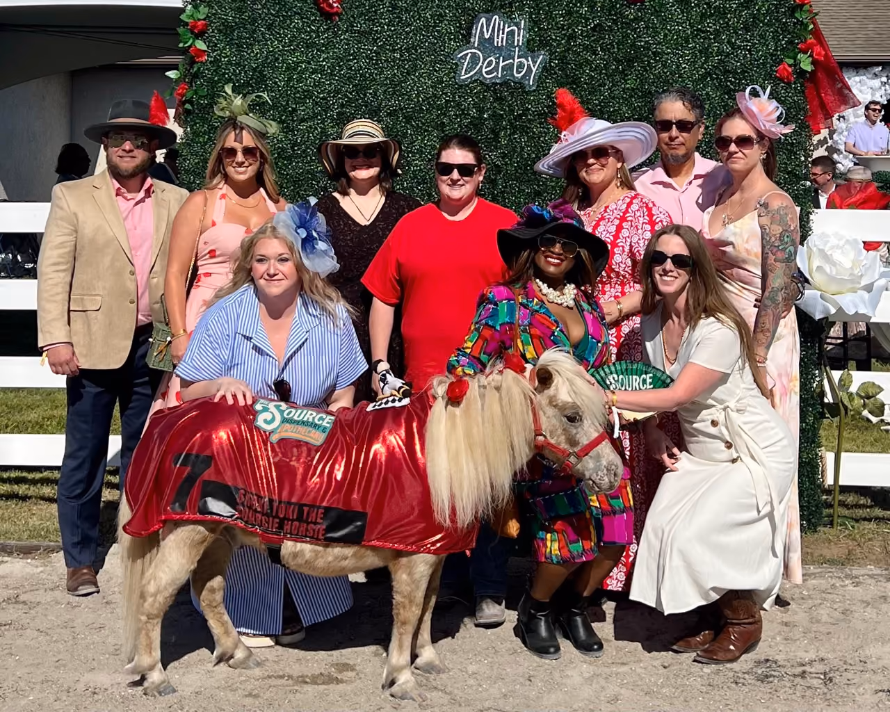 The Source team posing with Shoki Toki, 'The Sourcie Horsie,' a miniature horse wearing a red number 7 jockey blanket, in front of a floral 'Mini Derby' backdrop.