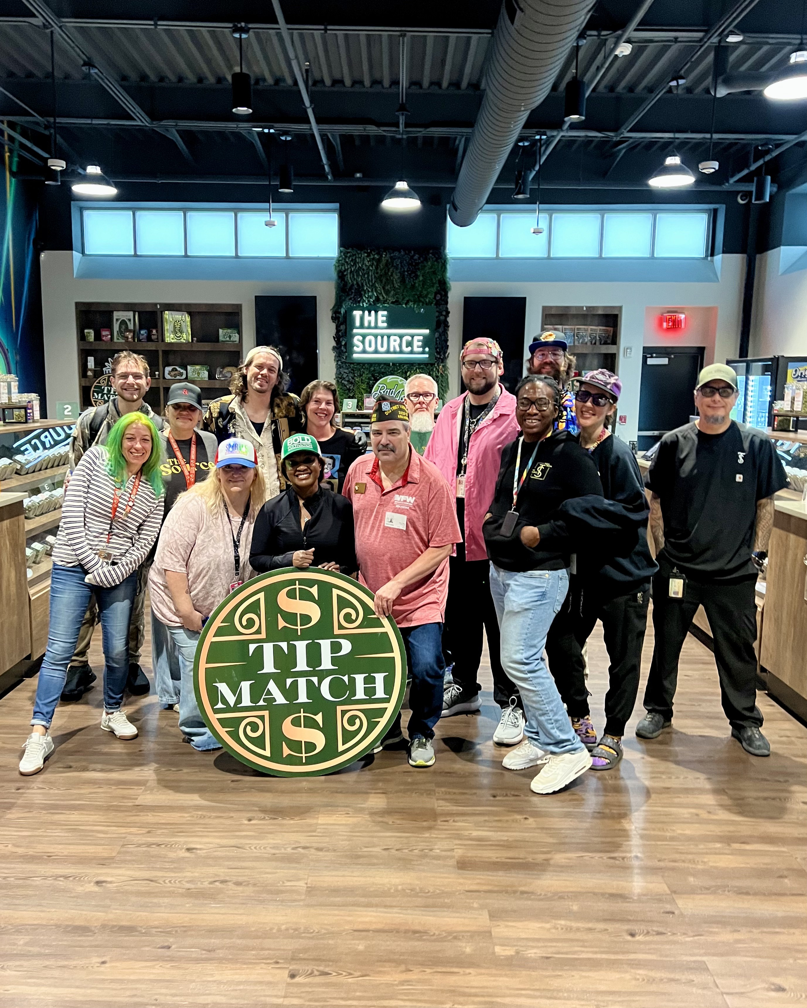 Large group photo for Tip Match with VFW Post 3031: thirteen team members posing together in the retail showroom holding the round green and gold Tip Match sign, with 'THE SOURCE' neon signage visible behind them.