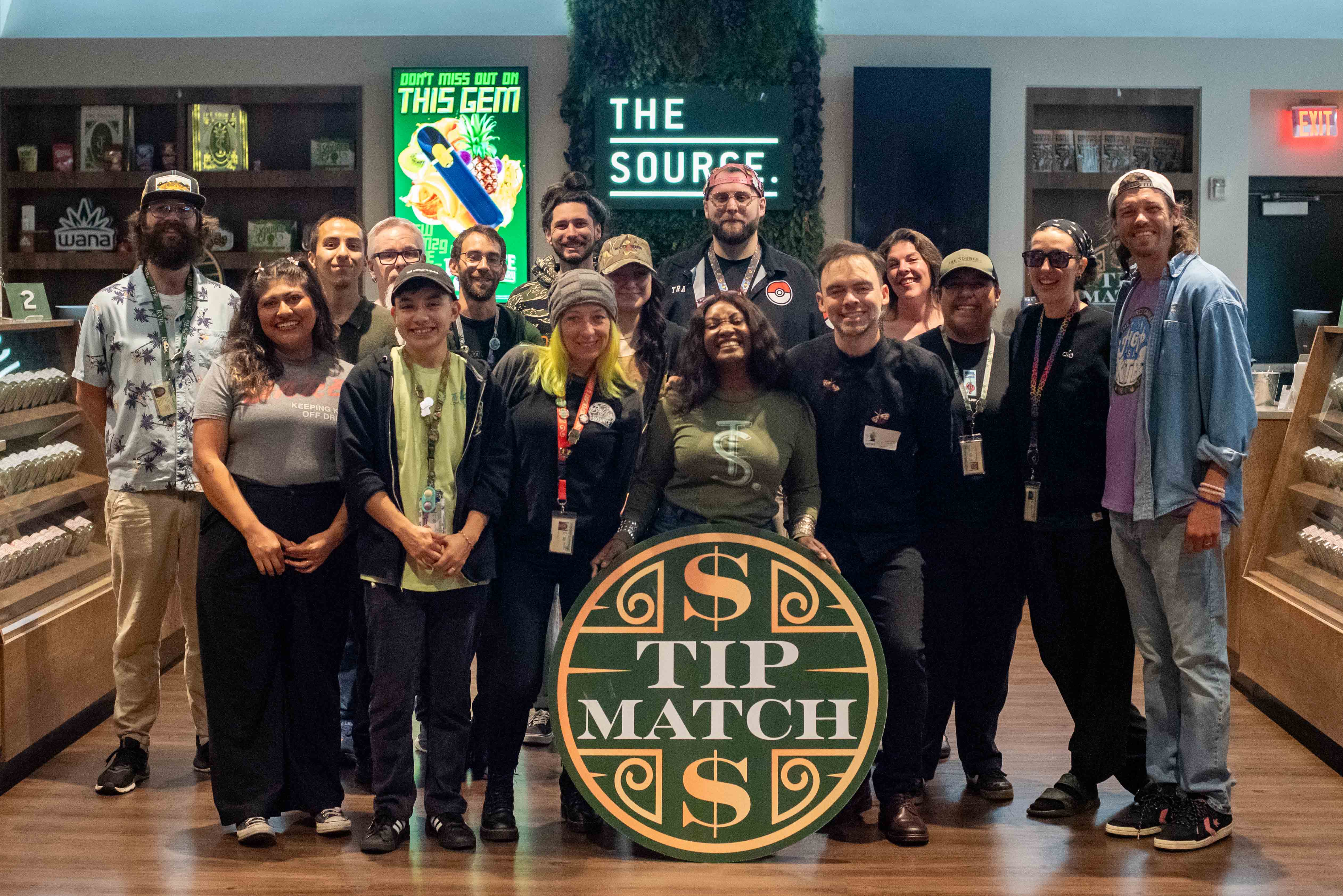 Tip Match with AID Arkansas: fourteen team members posing together in the retail showroom holding the round green and gold Tip Match sign, with 'THE SOURCE' signage and product displays visible behind them.