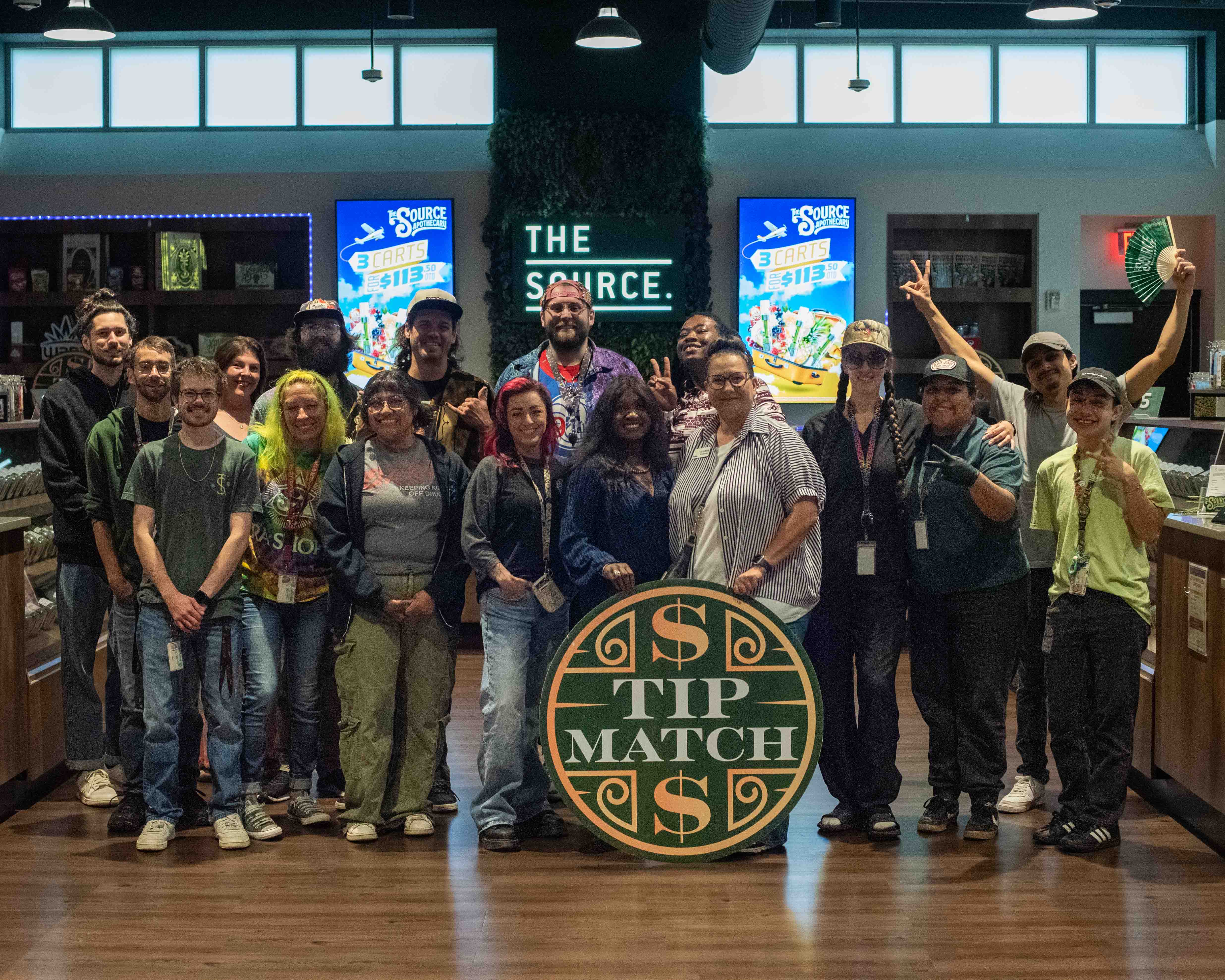 Tip Match with 7 Hills Homeless Center: fifteen Source team members and a 7 Hills team representative posing together in the retail showroom holding the round green and gold Tip Match sign, with 'THE SOURCE' signage and promotional screens visible.