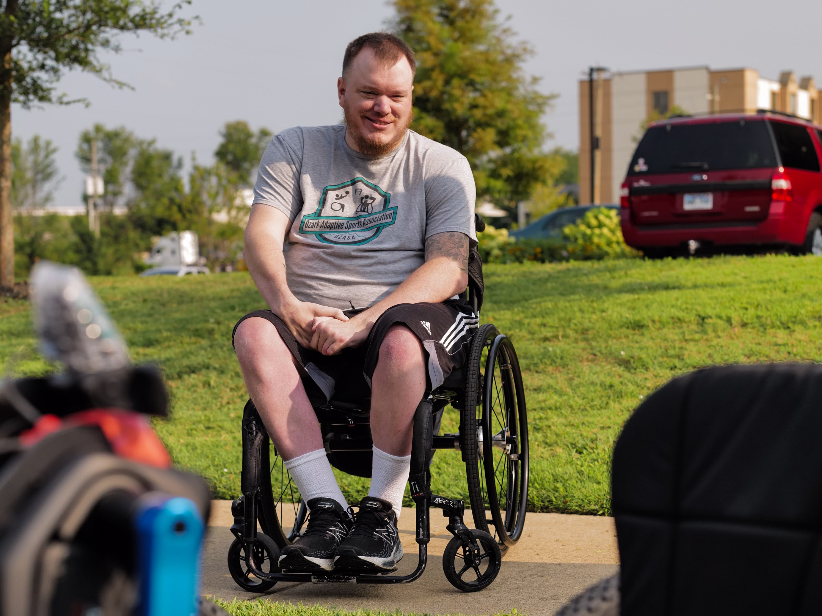 Young man in wheelchair smiling while wearing grey t-shirt with adaptive sports association logo, sitting outdoors on sunny day with green grass and buildings in background. He's wearing black Adidas shorts and athletic shoes, demonstrating inclusive sport