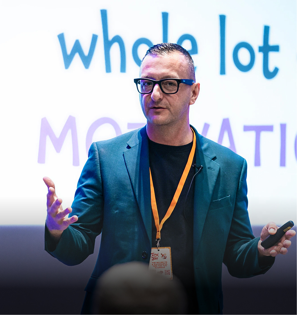 Speaker in glasses and teal jacket presenting with a remote, text 'whole lot' visible behind him.