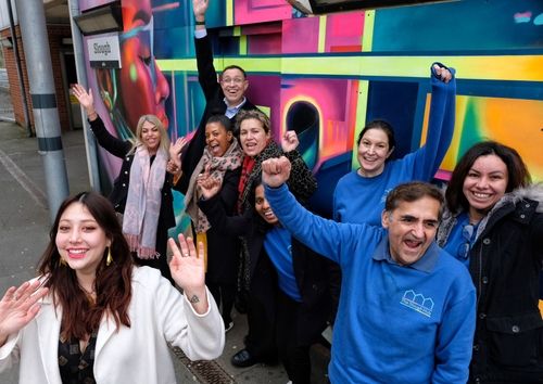 A cheerful group of individuals stands in front of a colourful mural, radiating happiness and excitement in a lively atmosphere.