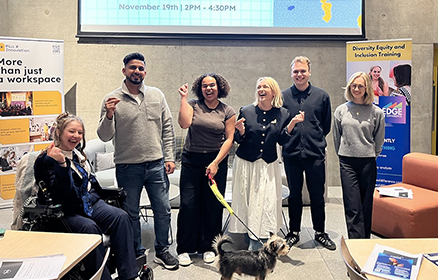 6 people (Calliope, Naqueeb, Kaia, Kerry, Sam and Alex) and Kaia’s dog, smiling and posing for a photo. They are stood in front of a giant Screen reading, “The Theory Of Change In Innovation” with a qr code to scan, with two branded banners - one on their left, the other on their right.