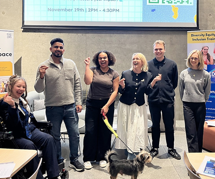 6 people (Calliope, Naqueeb, Kaia, Kerry, Sam and Alex) and Kaia’s dog, smiling and posing for a photo. They are stood in front of a giant Screen reading, “The Theory Of Change In Innovation” with a qr code to scan, with two branded banners - one on their left, the other on their right.