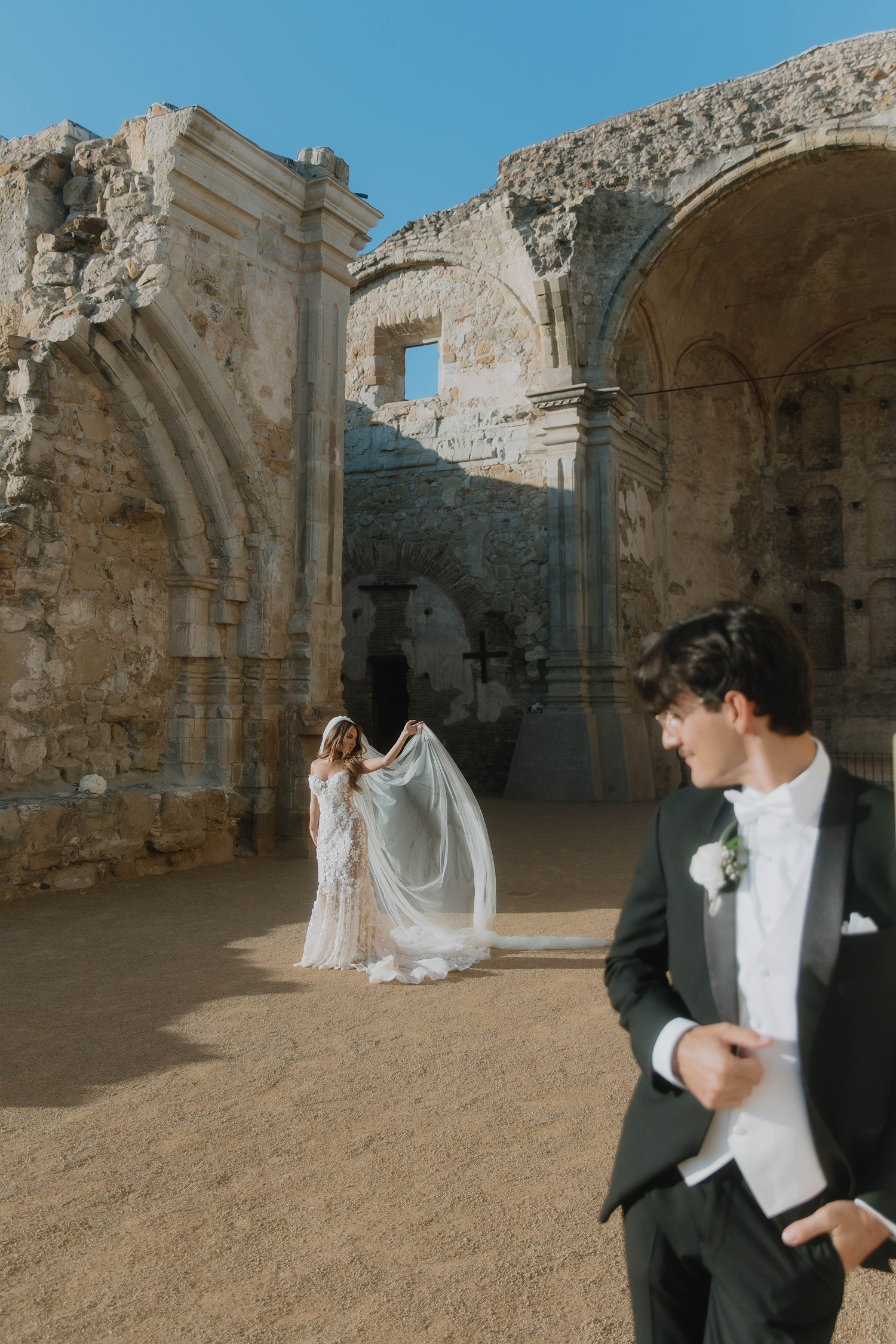 Bride in a white wedding gown holding her veil under ancient stone arches, groom in a black tuxedo looking towards her.