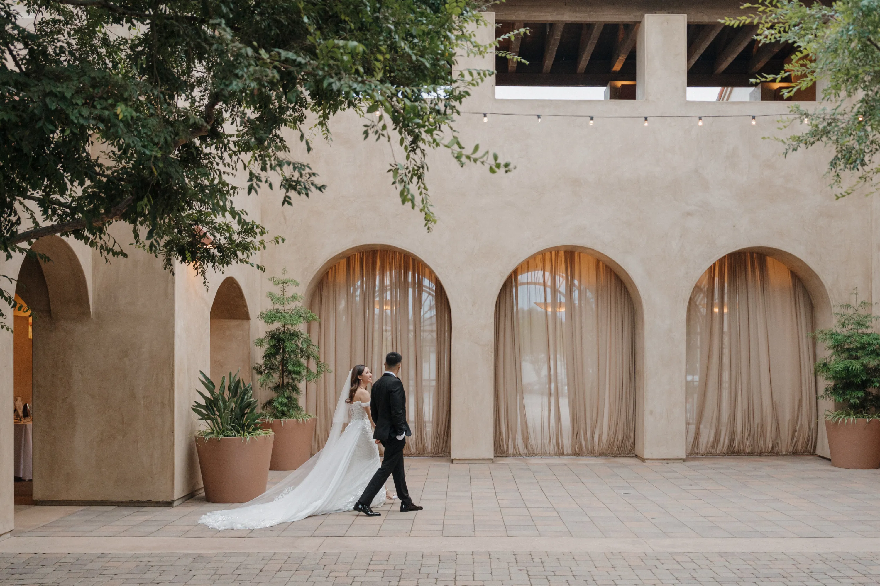 Bride in a white gown and groom in a black suit walking hand in hand by beige building with tall arched windows and potted plants.