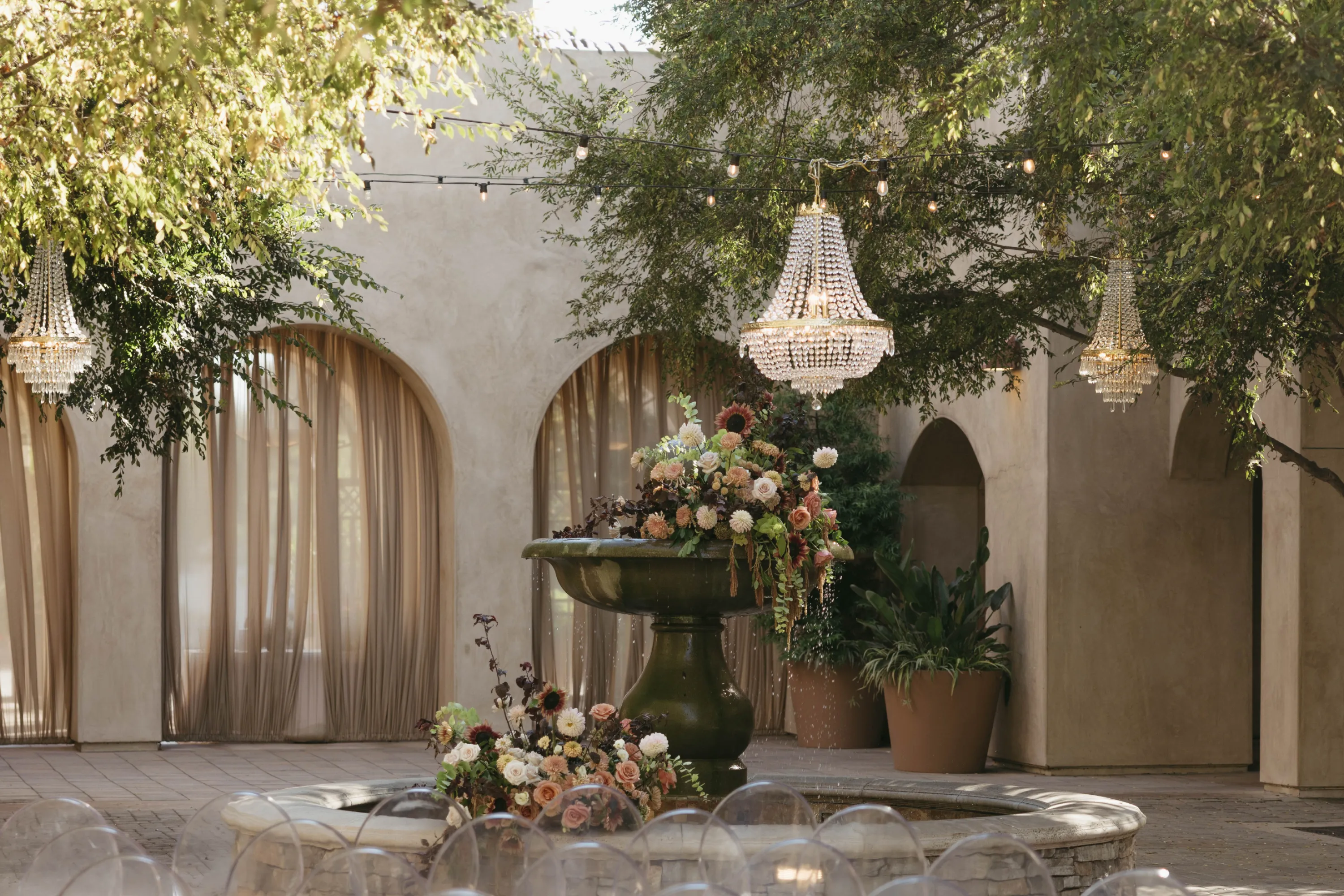 Outdoor courtyard with a central fountain decorated with floral arrangements, surrounded by arched windows with sheer curtains and hanging crystal chandeliers.