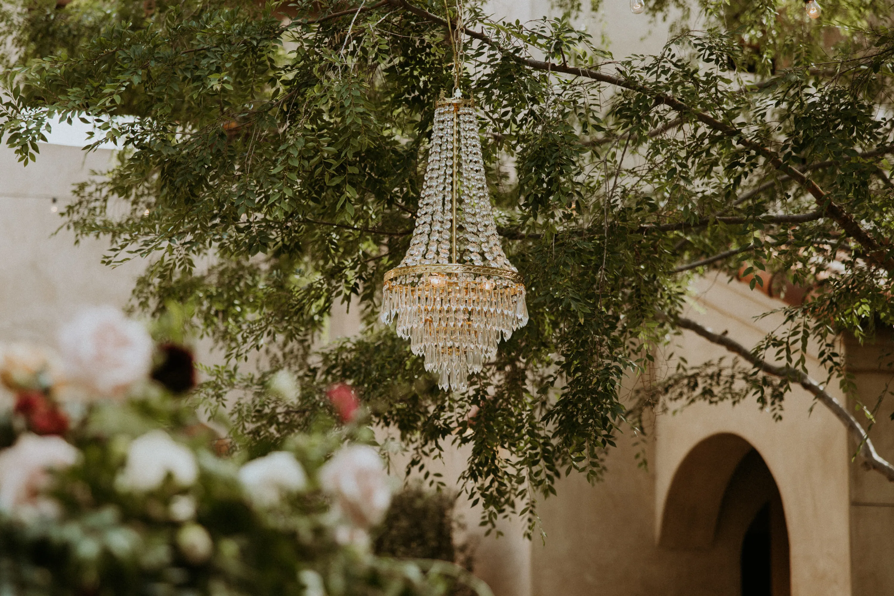 Crystal chandelier hanging from a tree branch with green leaves and blurred flowers in the foreground, set against a beige building with an arched doorway.