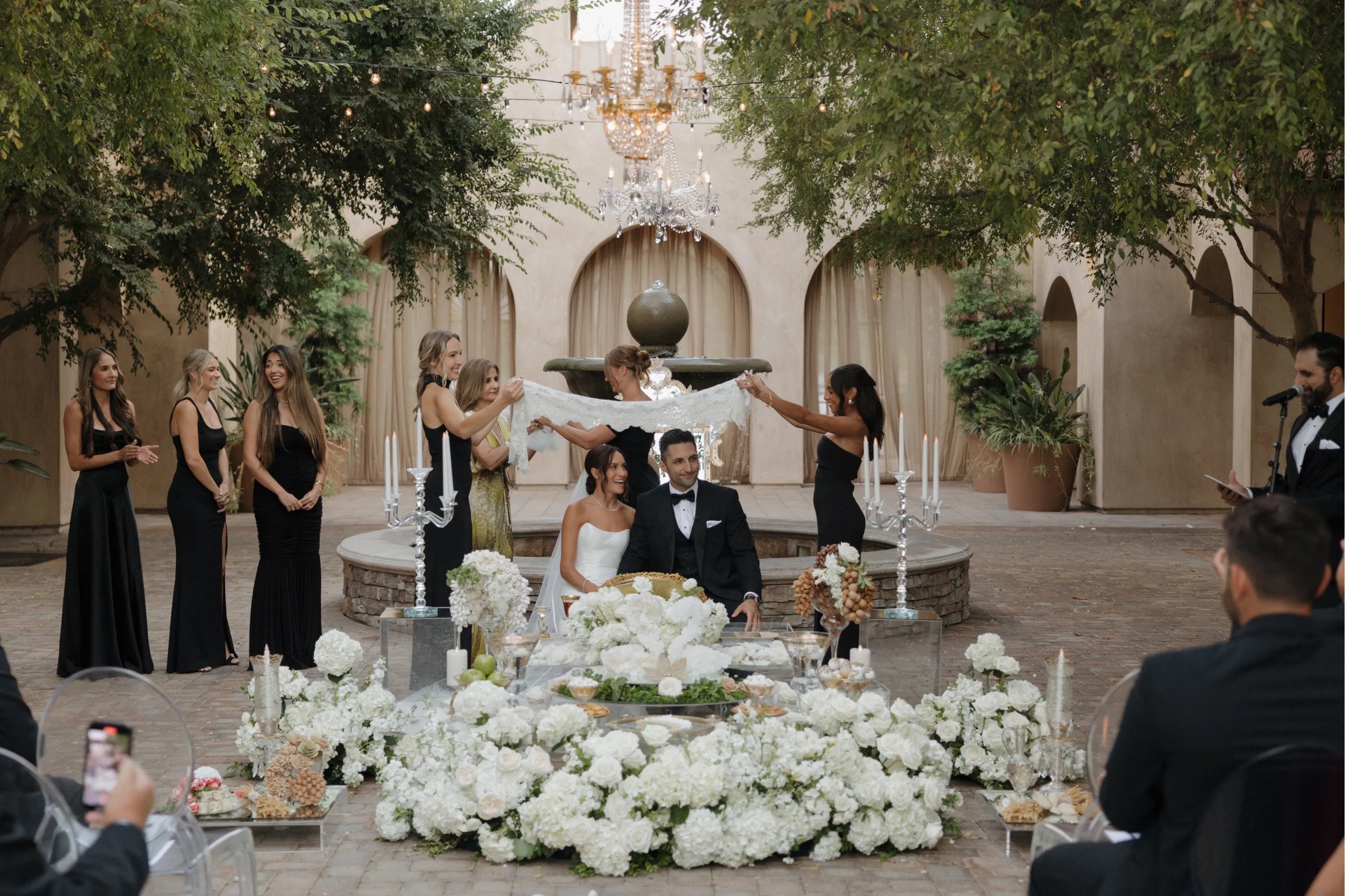 Bride and groom seated in front of a floral arrangement while bridesmaids hold a white cloth over them during an outdoor wedding ceremony.