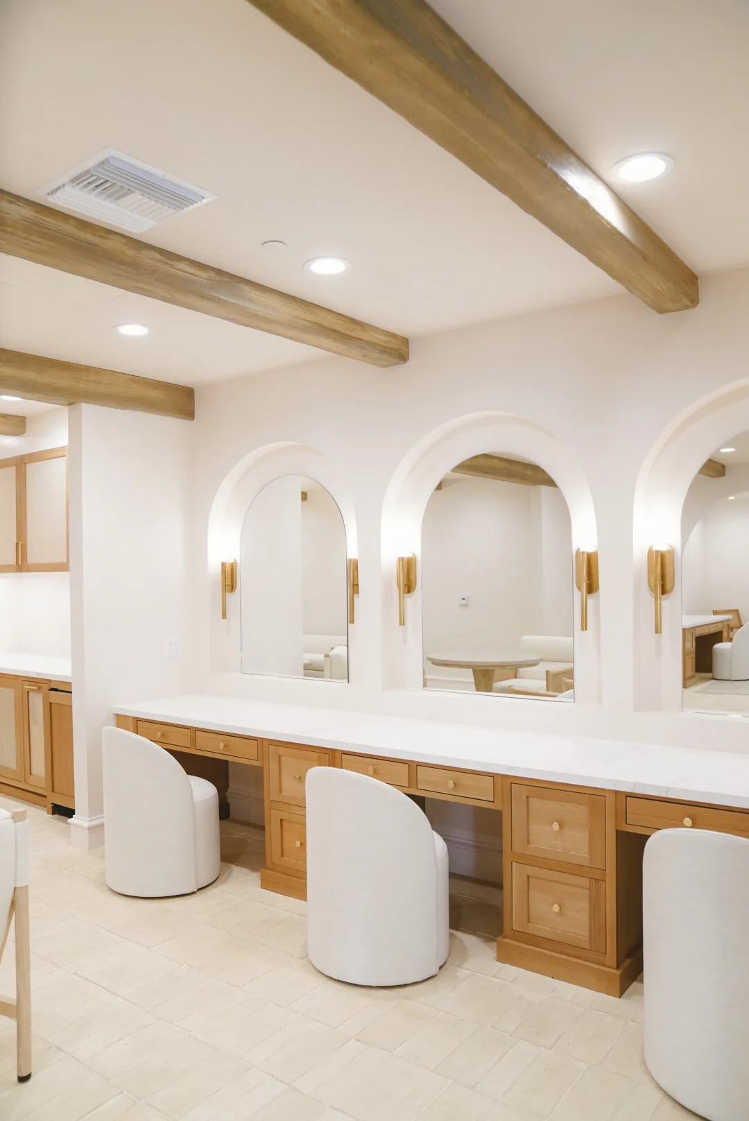 Modern dressing room featuring wooden vanity with drawers, white cushioned chairs, arched mirrors, and exposed ceiling beams.